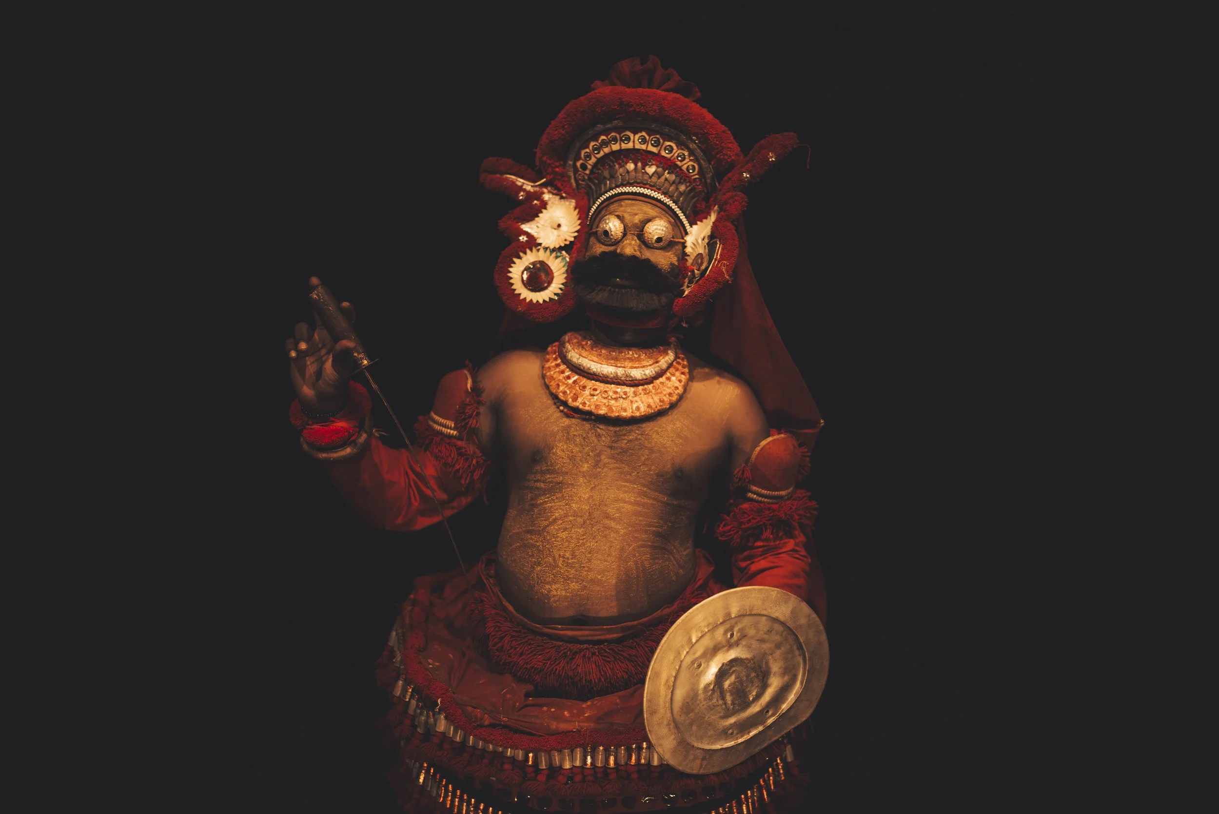 Traditional Indian dancer dressed in vibrant costume with a decorative headdress, face mask, and holding a shield, performing against a dark background.