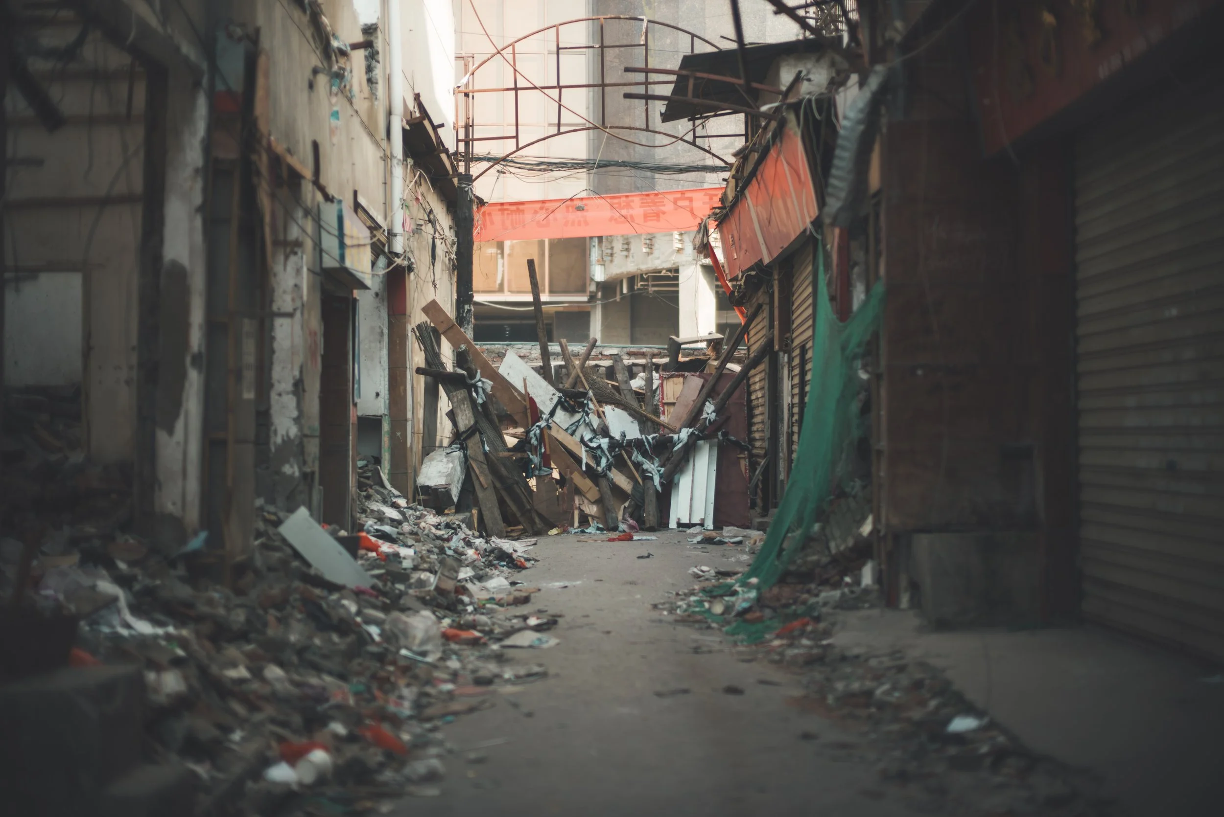 A rundown alleyway filled with rubble, debris, and broken wooden structures, with old buildings on either side and a red banner hanging across the alley.