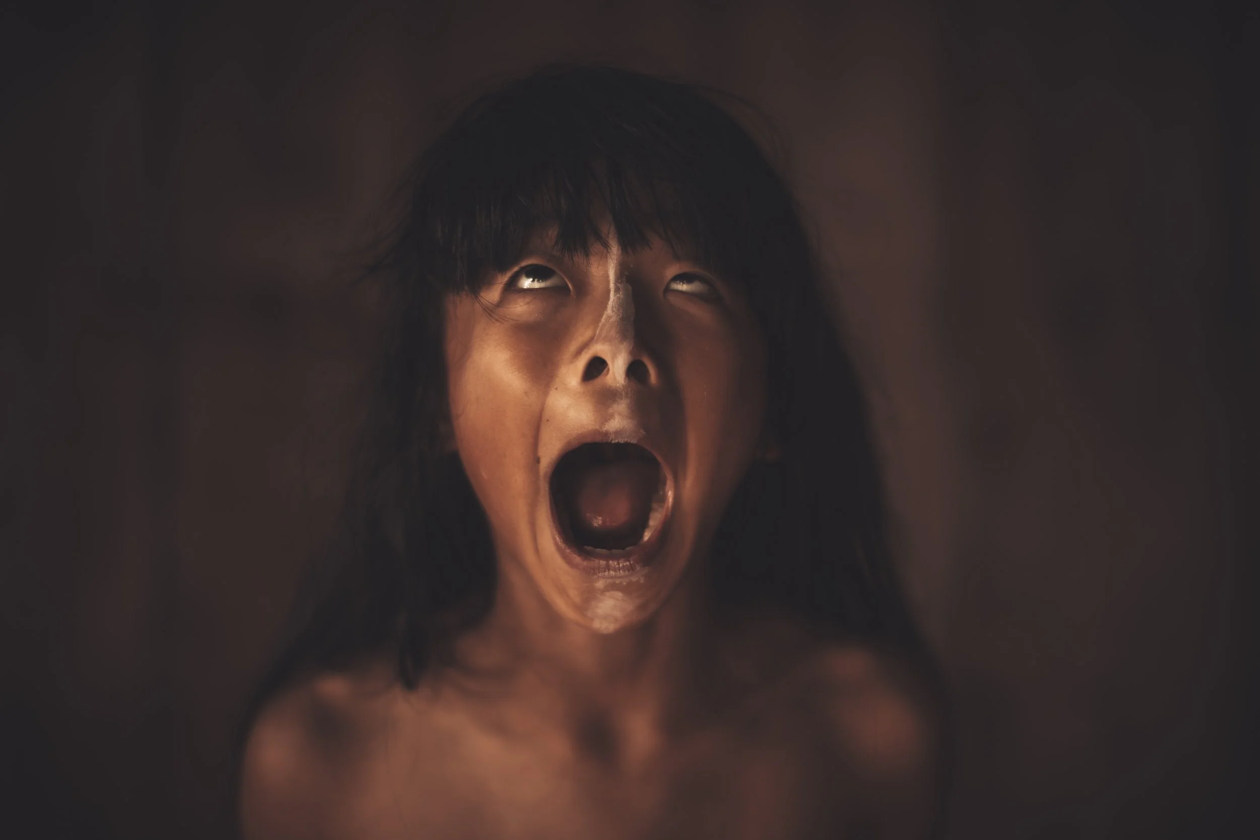 A woman with long dark hair and brown skin, screaming with her mouth wide open and eyes looking upward.