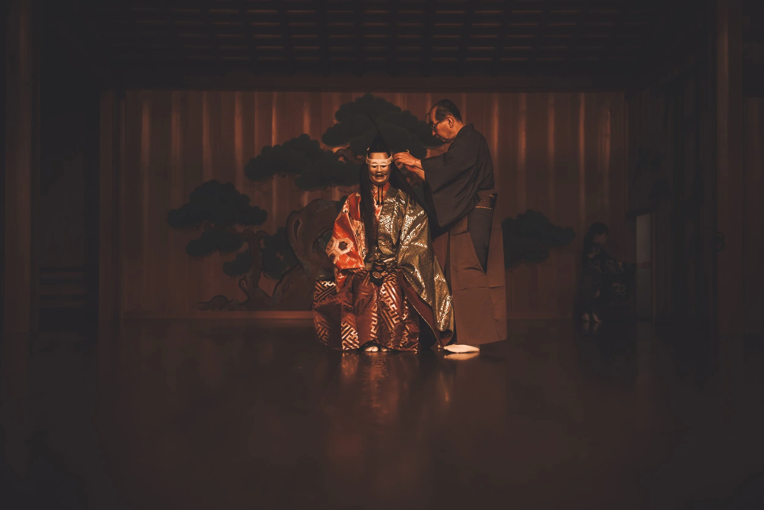 A person in traditional Japanese kabuki theater makeup and clothing is seated on stage, with another person standing beside them adjusting their costume, while a painted backdrop of a pine tree is visible behind them.