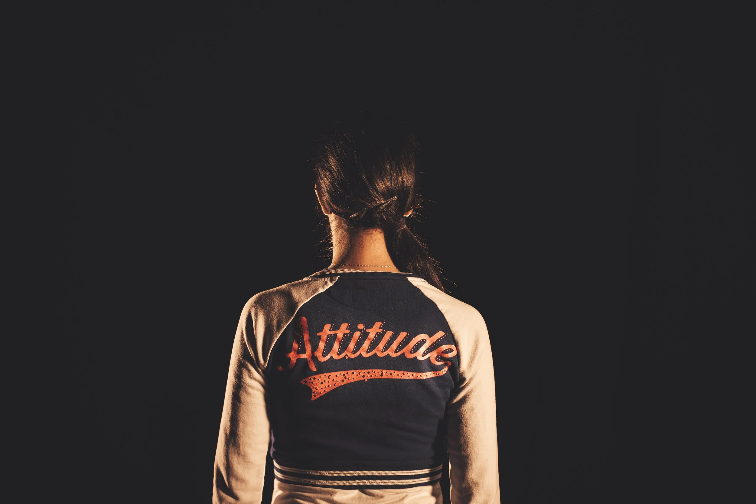 Back of a person wearing a sports jersey with the words 'Attitude' in orange text, standing against a dark background.