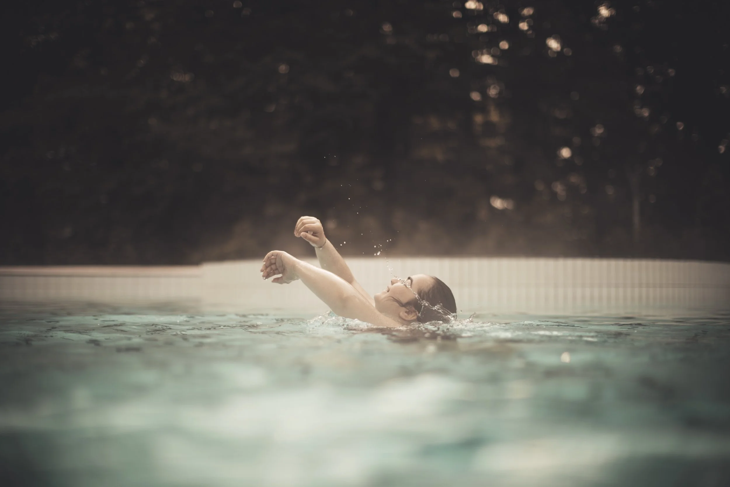 A woman swimming in a pool, with her head above water, wearing goggles and a swimsuit, in an indoor pool with a dark background.