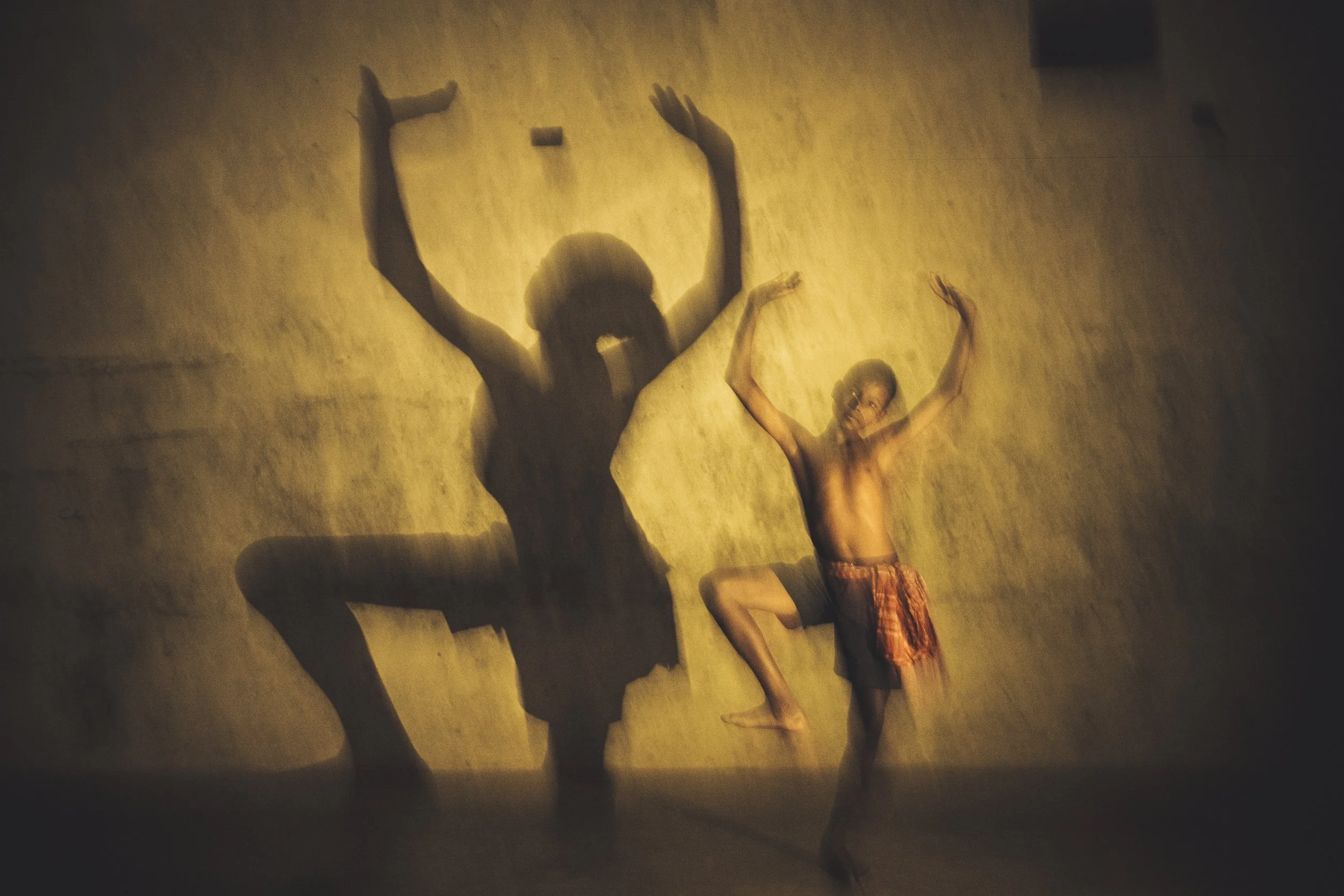 A person sitting on the floor with legs crossed and arms raised, casting a shadow on a wall. The wall is illuminated with warm lighting, creating a silhouette effect.