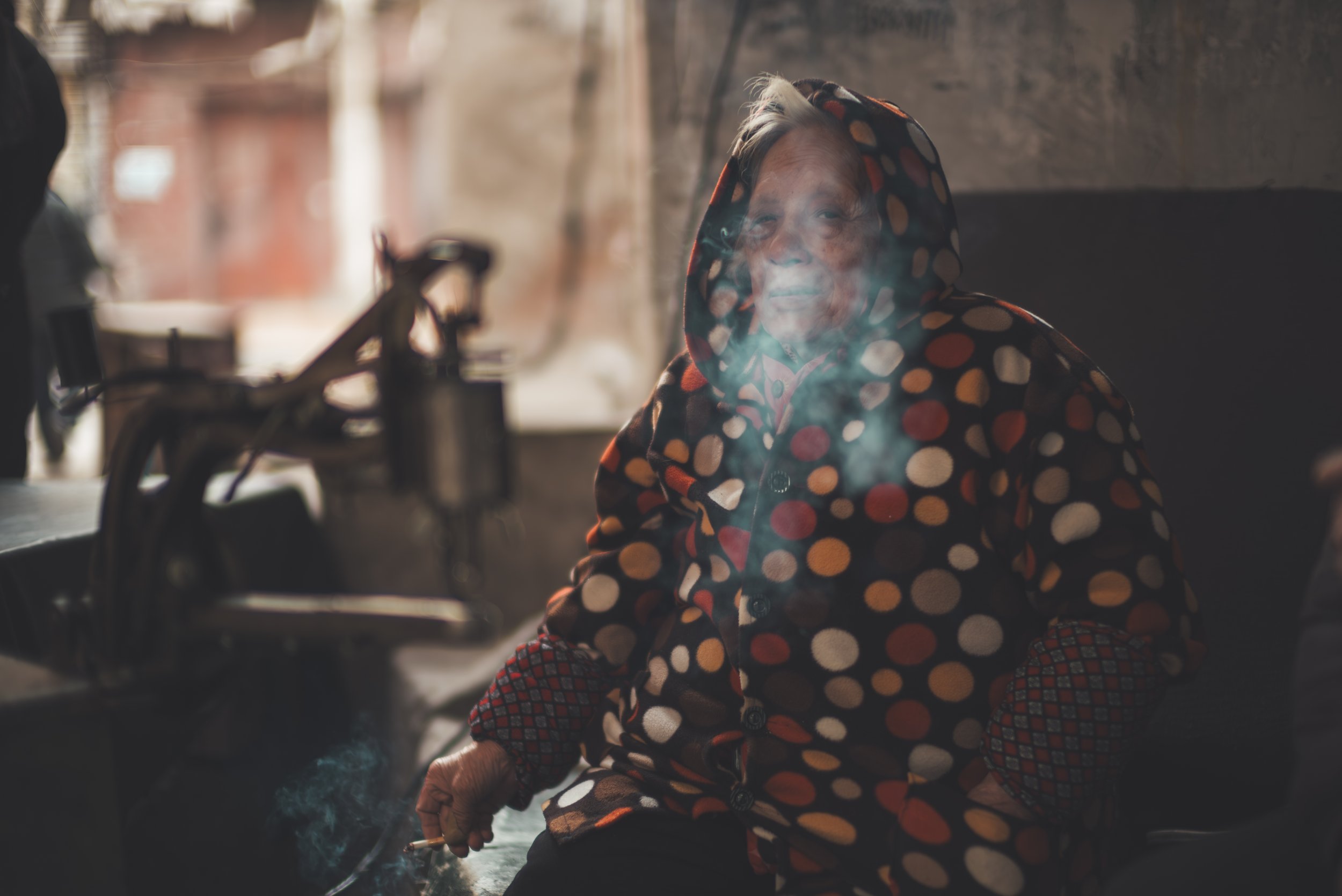 An older woman wearing a polka dot hoodie, smoking, sitting inside a dimly lit room with a bicycle in the background.