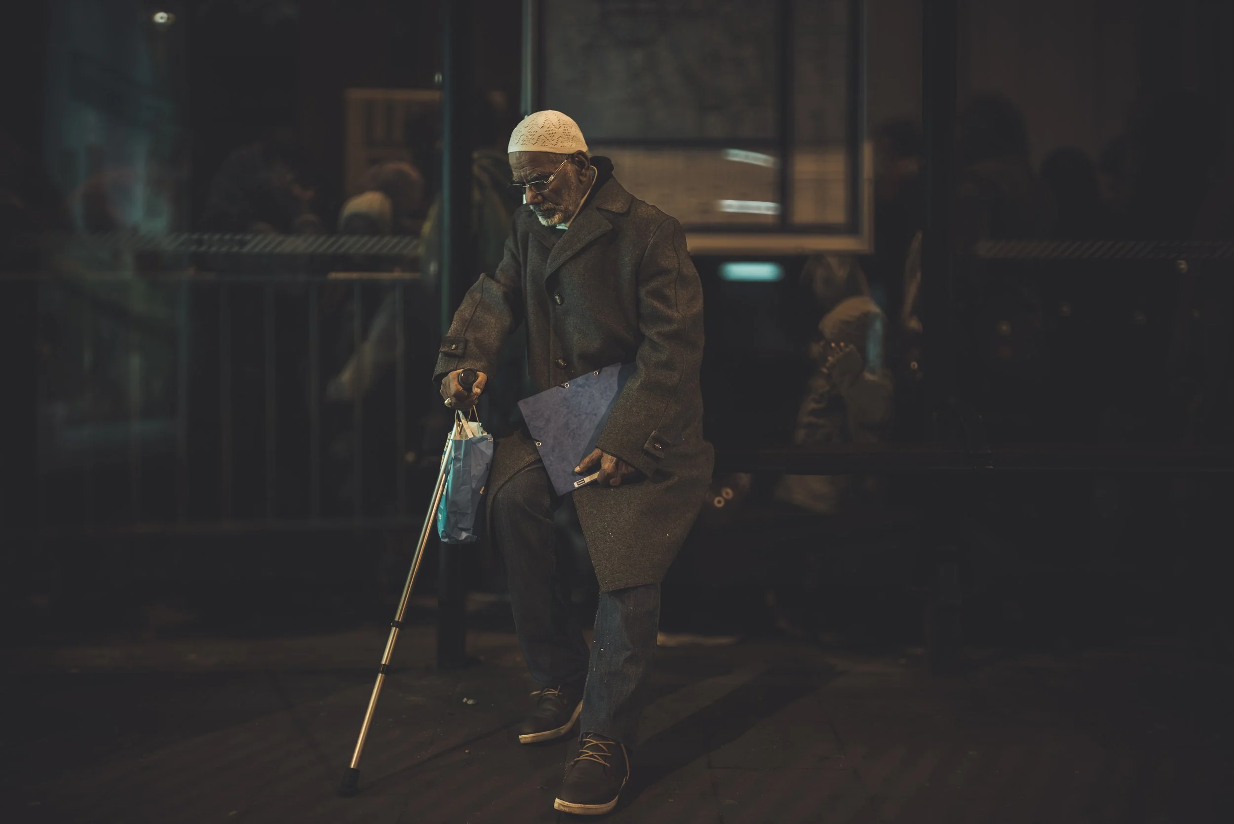 An elderly man wearing a white knit cap, glasses, a gray overcoat, and dark pants walks with a cane through a dimly lit area at night, carrying shopping bags.
