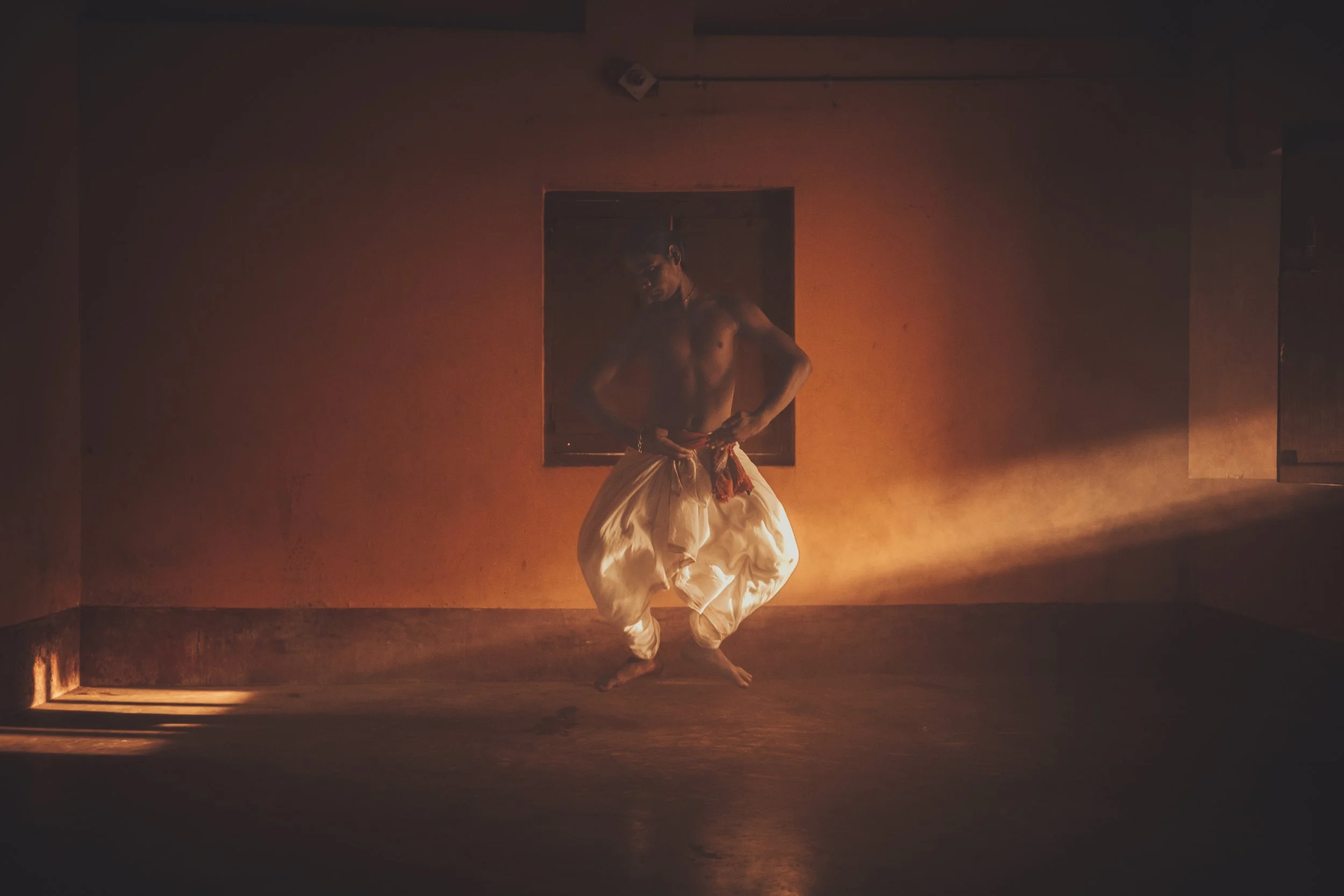 A shirtless man wearing traditional white pants is dancing in a dimly lit room with orange lighting, with a small window in the background.