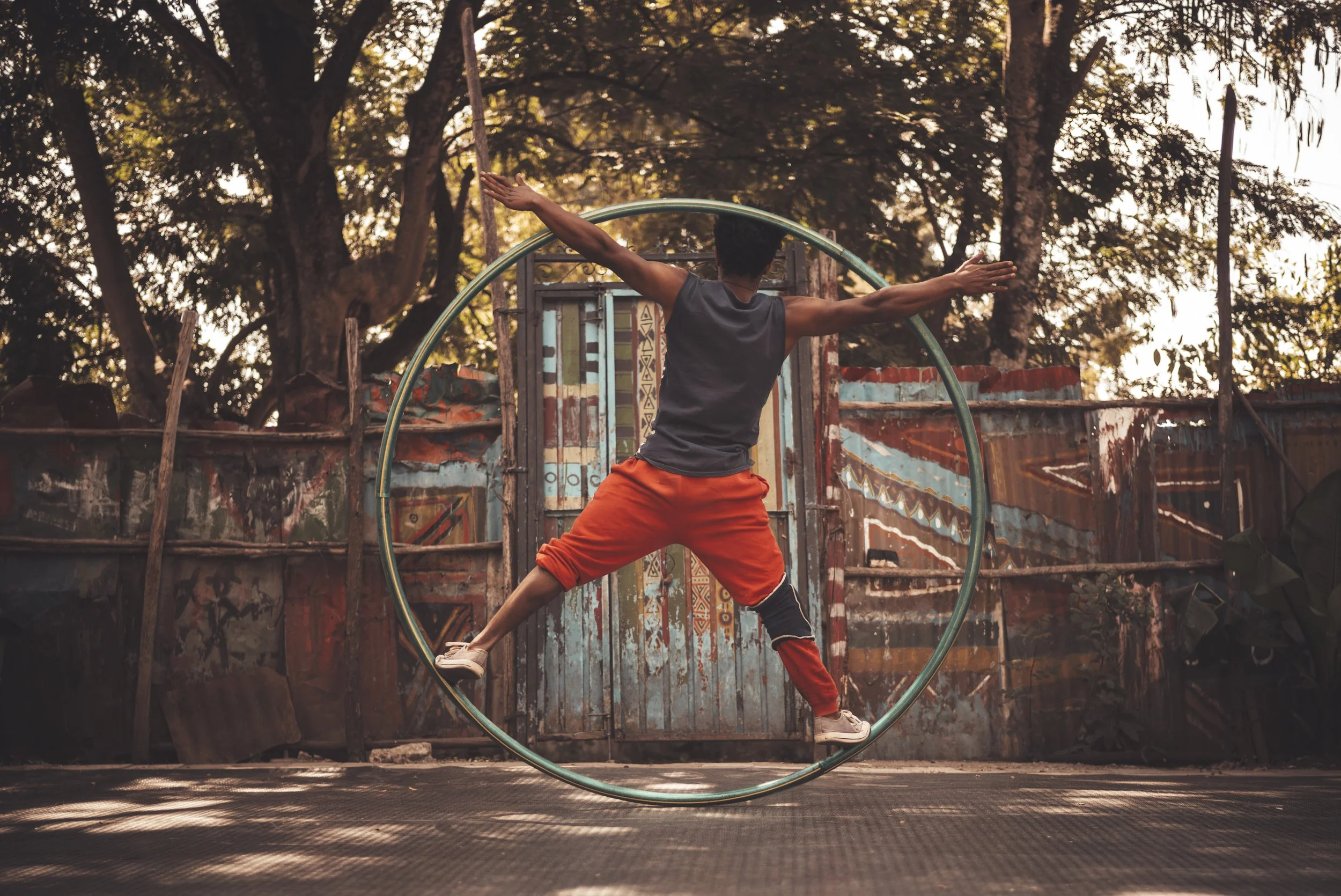 Young person performing a balance move on a large hula hoop outdoors during sunset, with trees and a decorated fence in the background.