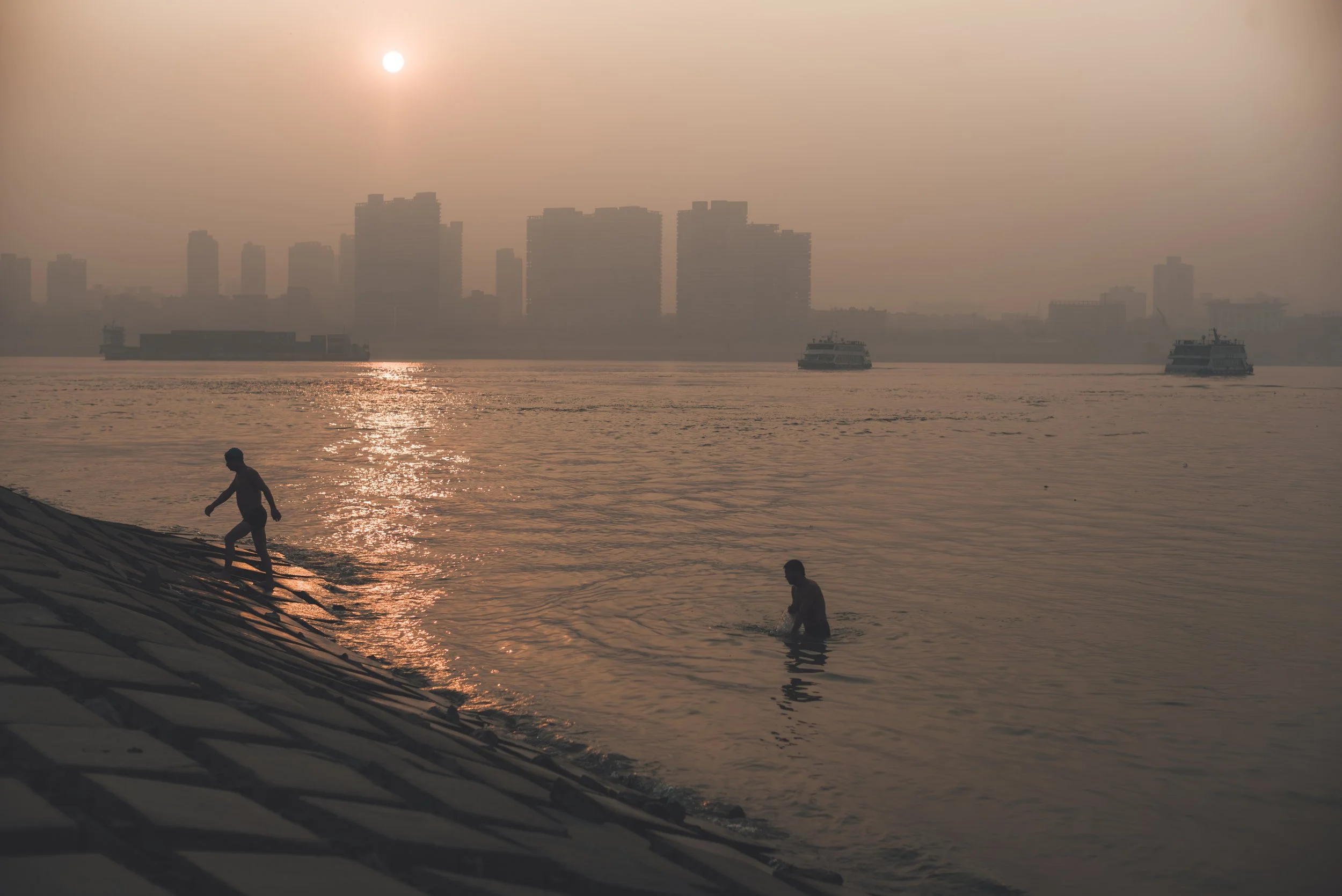 People playing and swimming in the water at the shoreline during sunset, with a city skyline and boats in the background.