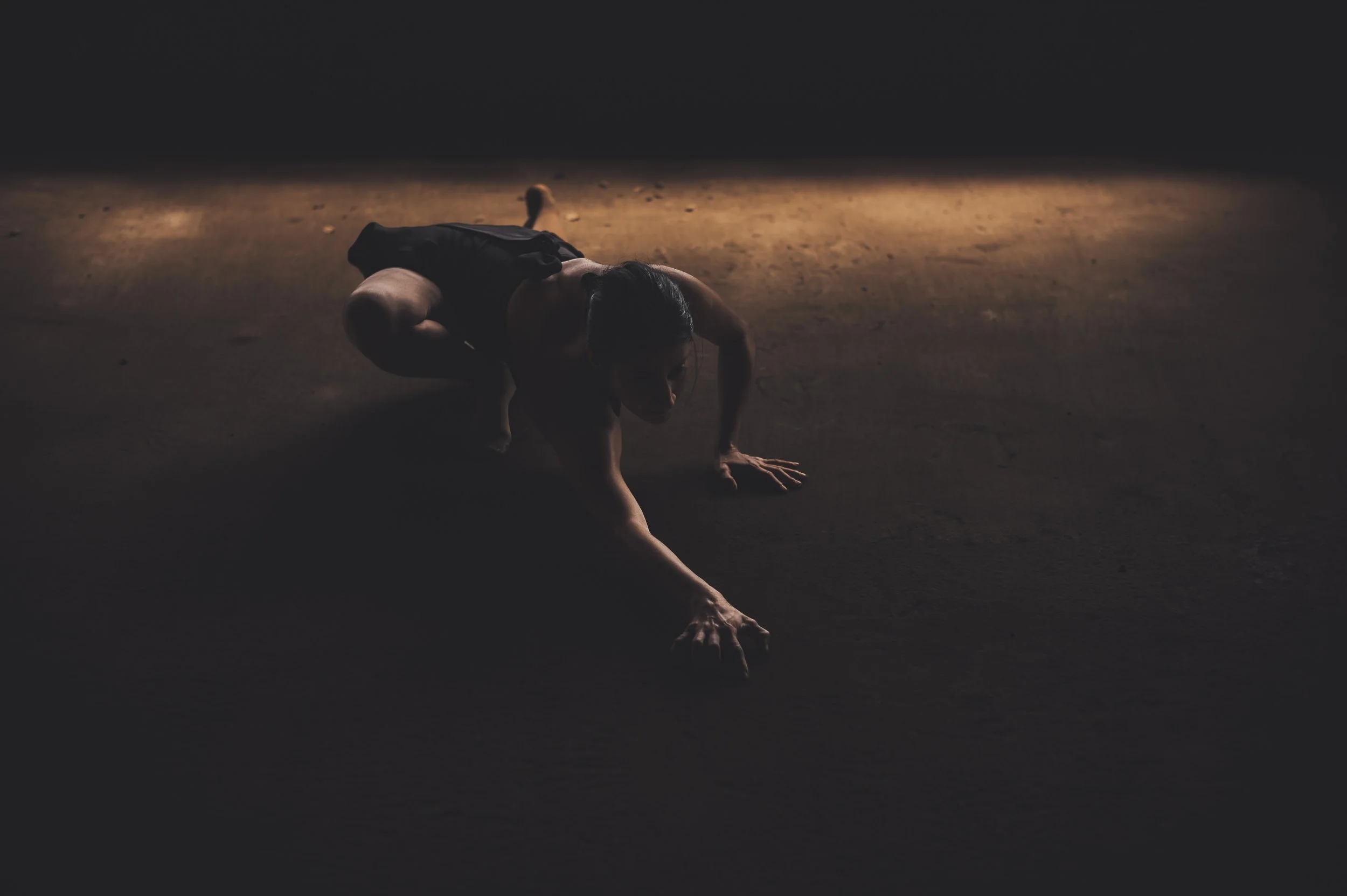 A person is crawling on the ground in a dimly lit environment with a dark background, wearing dark clothing.