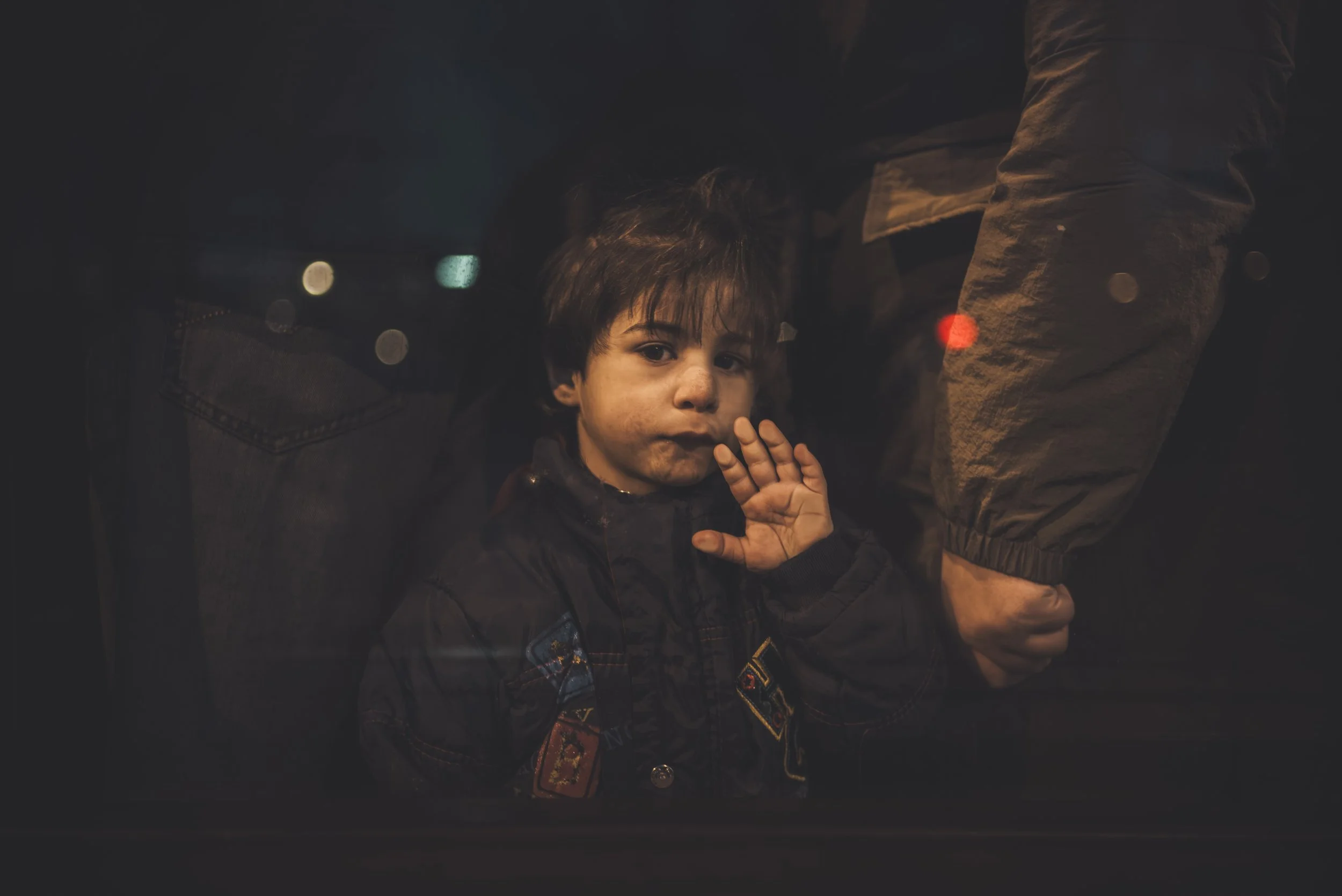 A young boy with dark hair and a dark jacket, with a dirty face, is standing between people at night. One person next to him has a jacket with a patch featuring an eagle. The boy's hand is raised near his face. The background shows blurry lights.