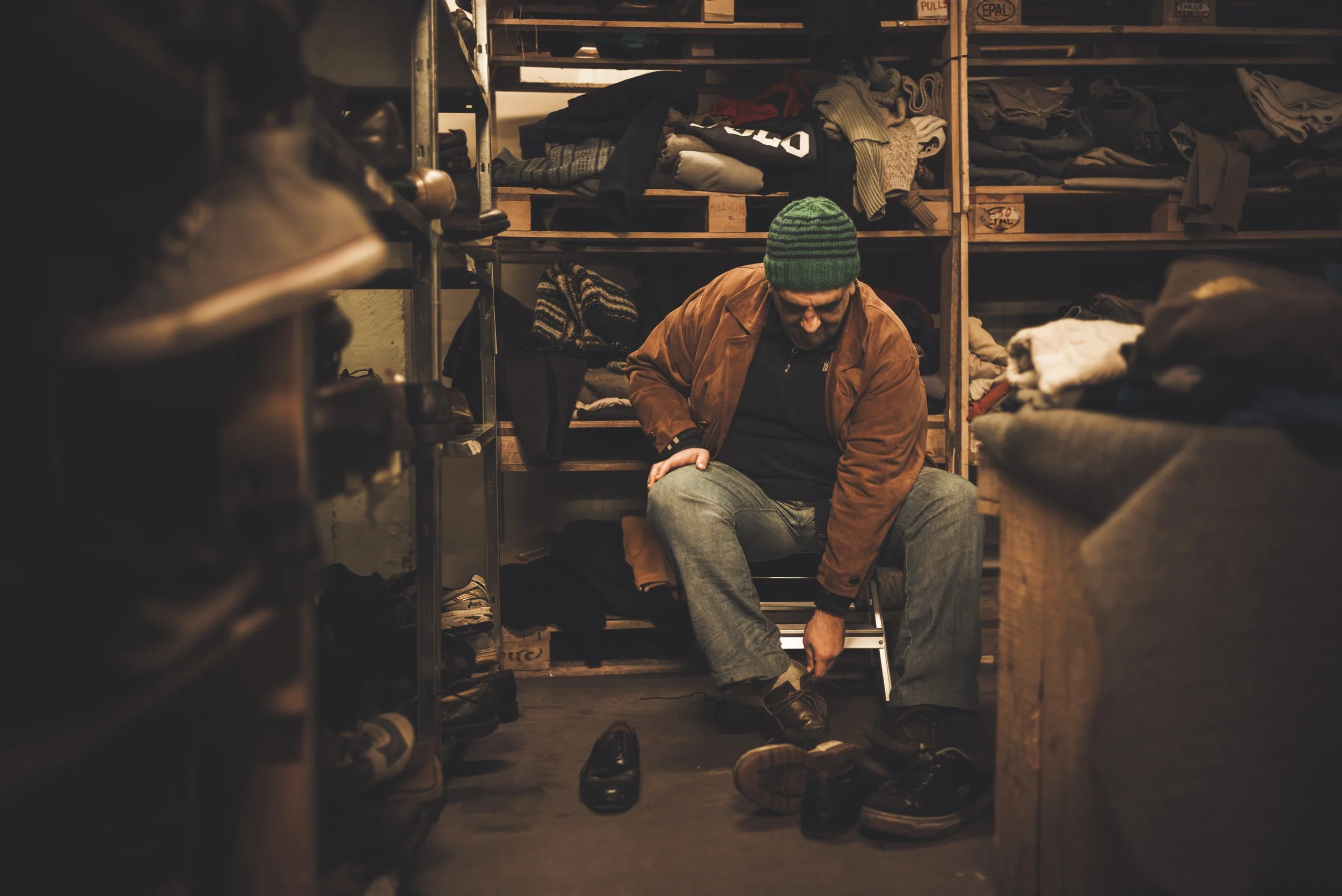 A man sitting in a shoe store, tying his shoe, surrounded by shelves filled with shoes and clothing.