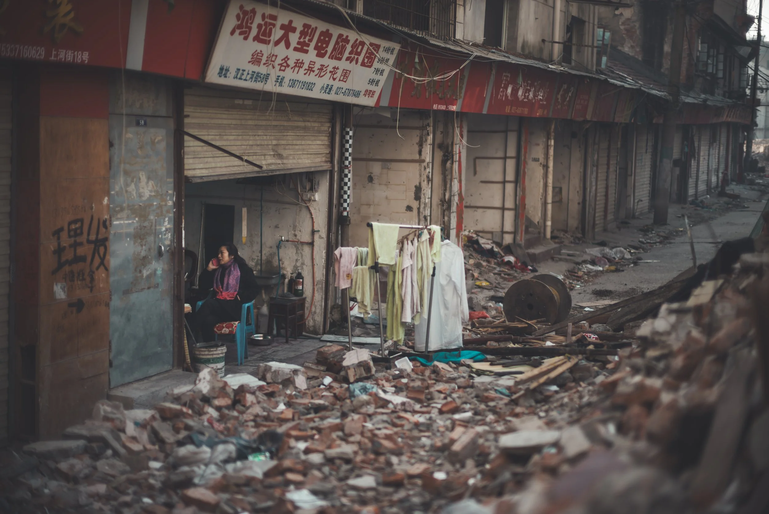 A woman sitting on a blue chair talking on a mobile phone outside a damaged building with rubble and debris on the ground, in an urban area with closed shop shutters.