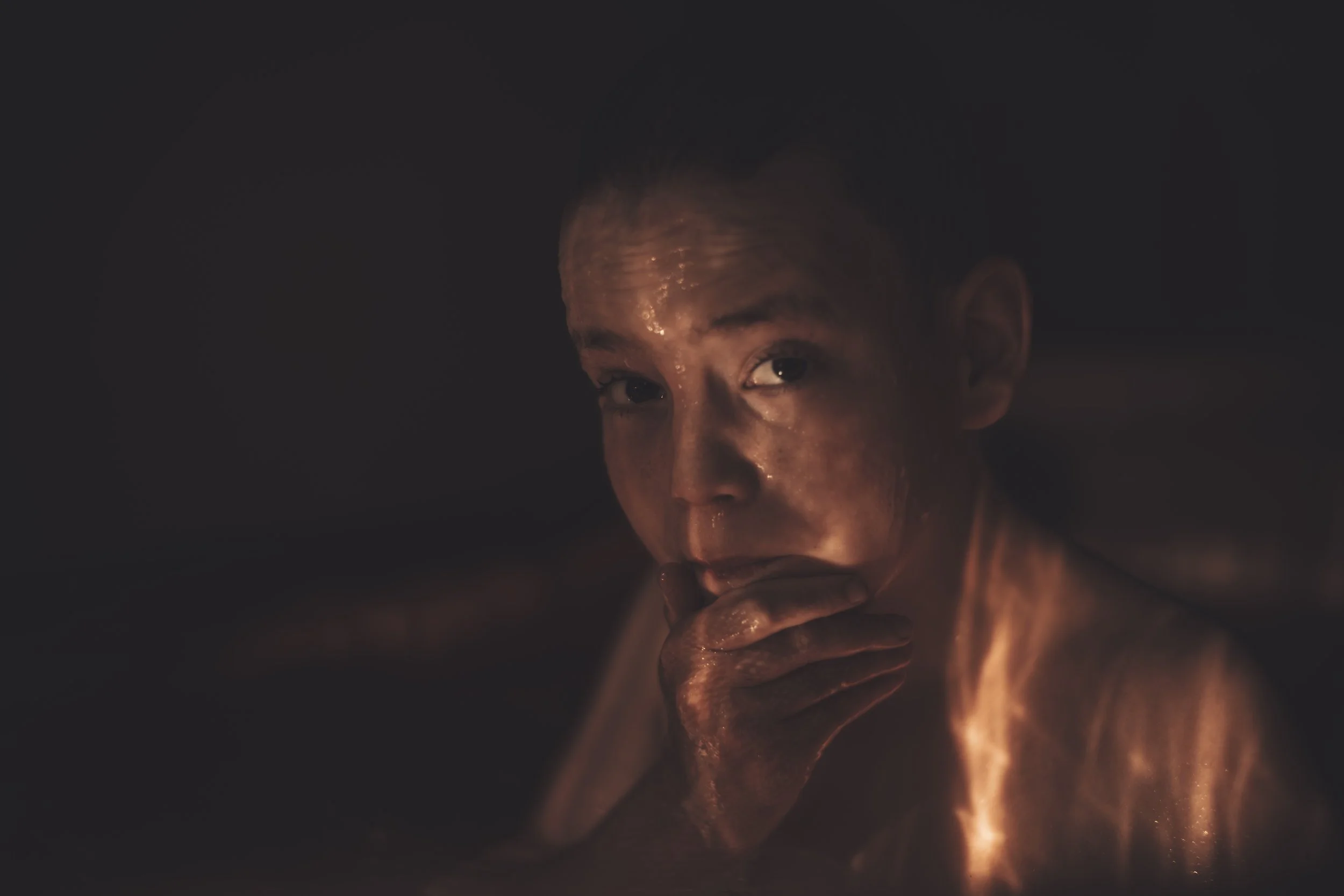 A woman with wet hair and skin is drinking from a glass in a dimly lit environment, illuminated by warm, orange-toned light.