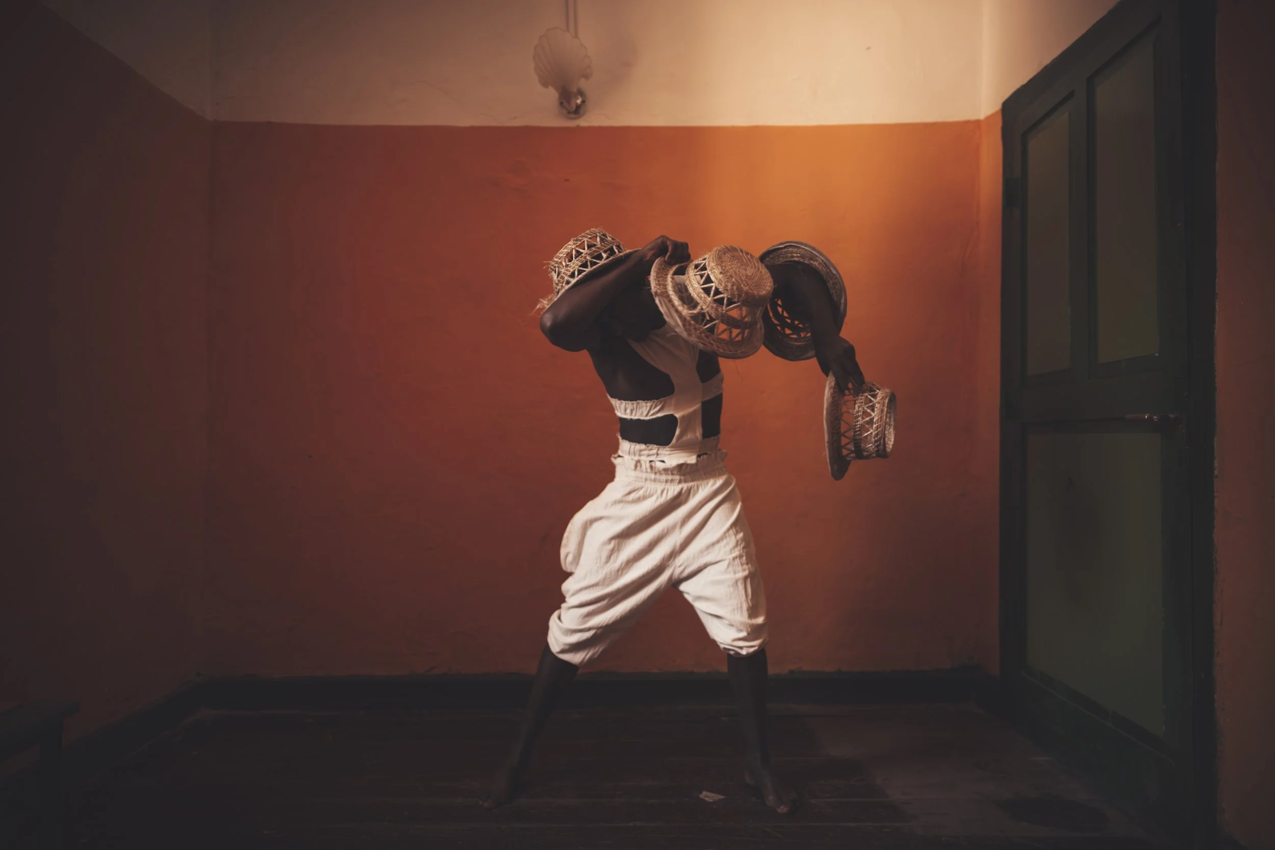 A person with dark skin dancing with traditional woven baskets on their head in a dimly lit room with orange and beige walls.