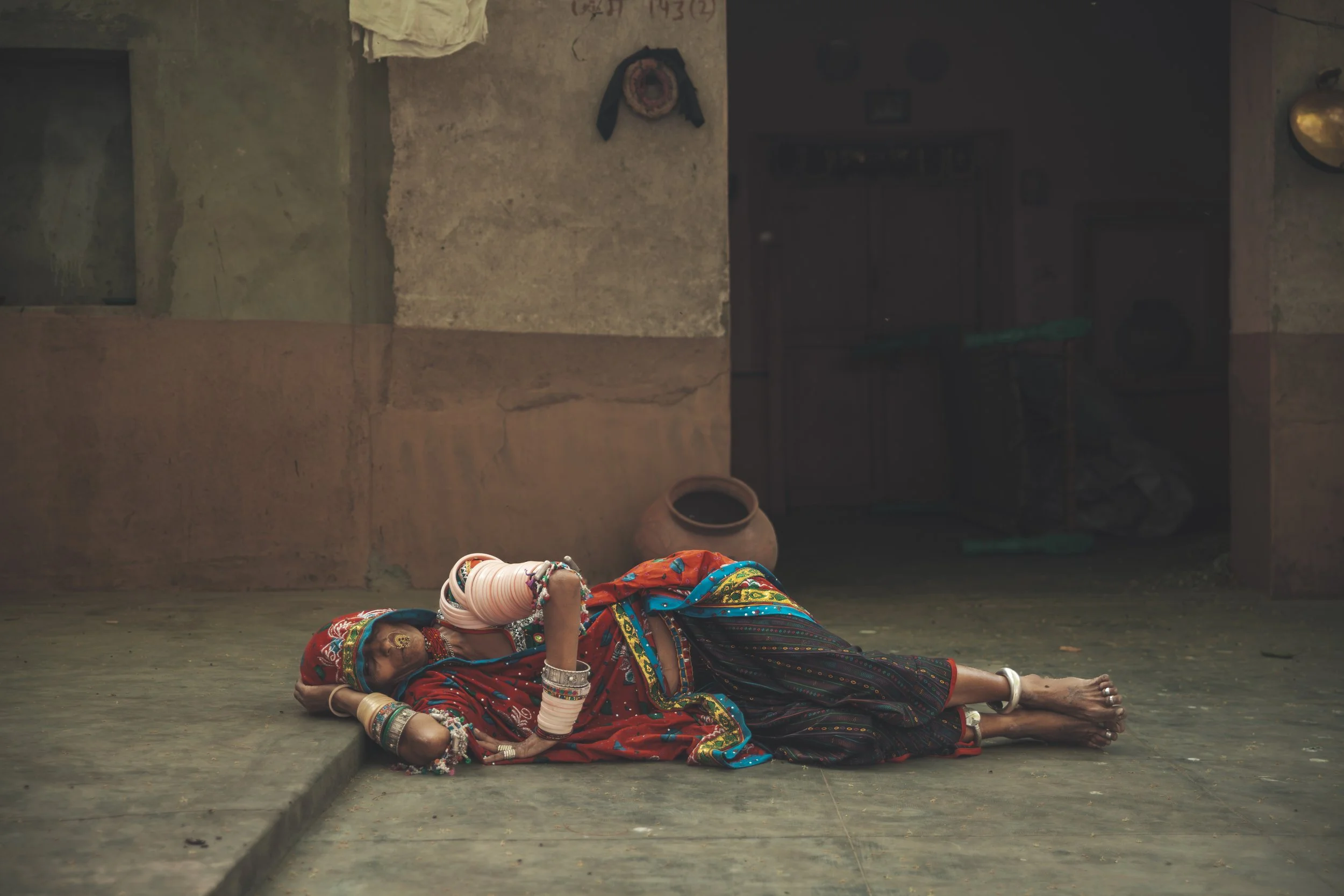 A woman dressed in colorful traditional Indian clothing and jewelry is lying on the ground in a doorway, appearing to be resting or unwell.