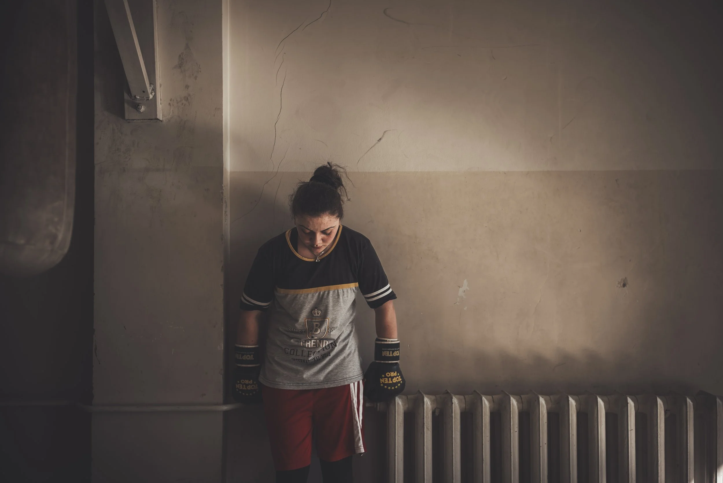 A young woman with dark hair tied up in a bun is wearing black boxing gloves and red shorts, standing against a plain, beige wall with cracked paint. She appears to be in a boxing or training stance, with her head slightly bowed.