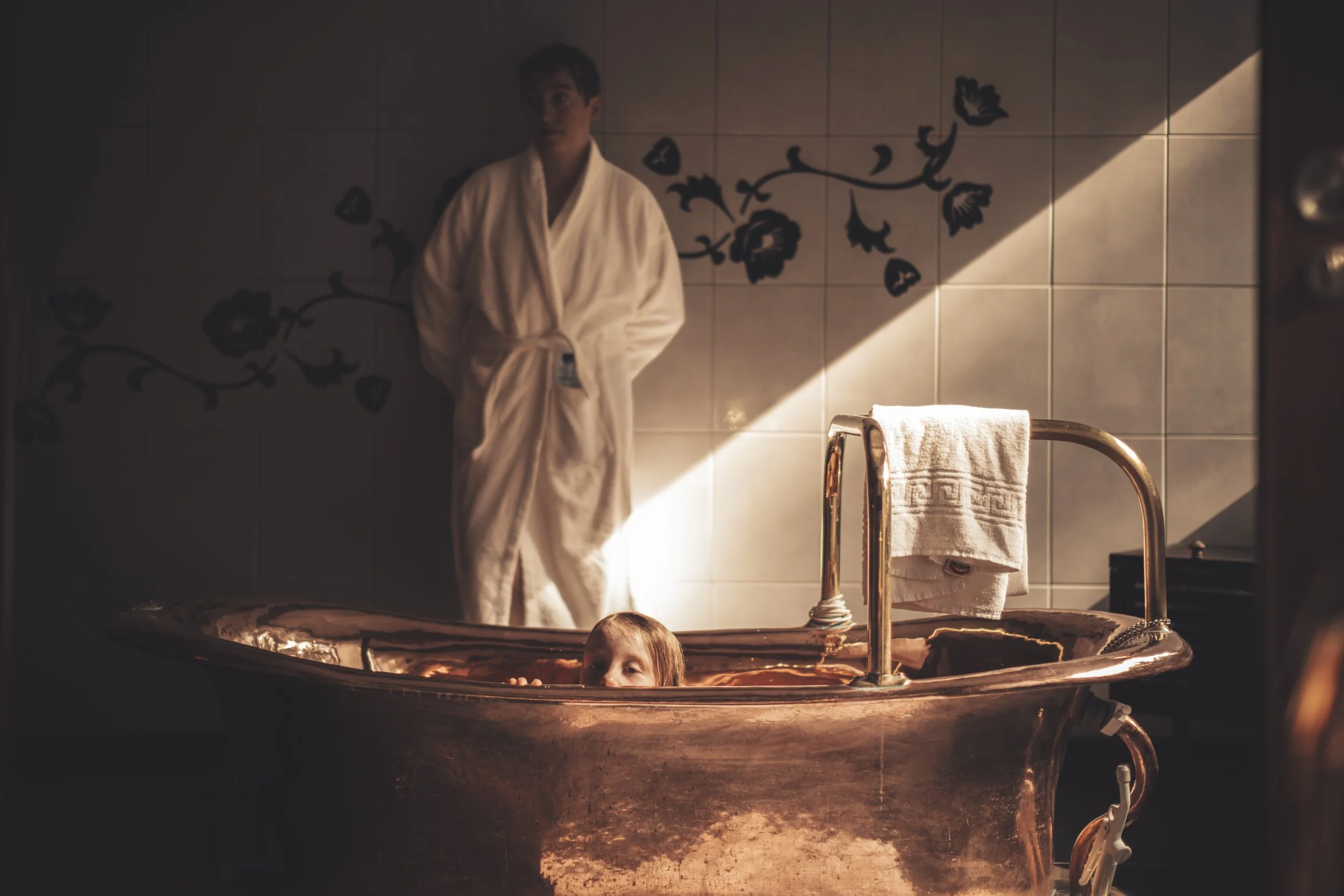 A young boy is submerged in a copper hot tub with only his head visible. A woman, possibly his mother, is standing behind the tub wearing a white bathrobe, leaning against a tiled wall. The scene is dimly lit, creating a cozy or intimate atmosphere.