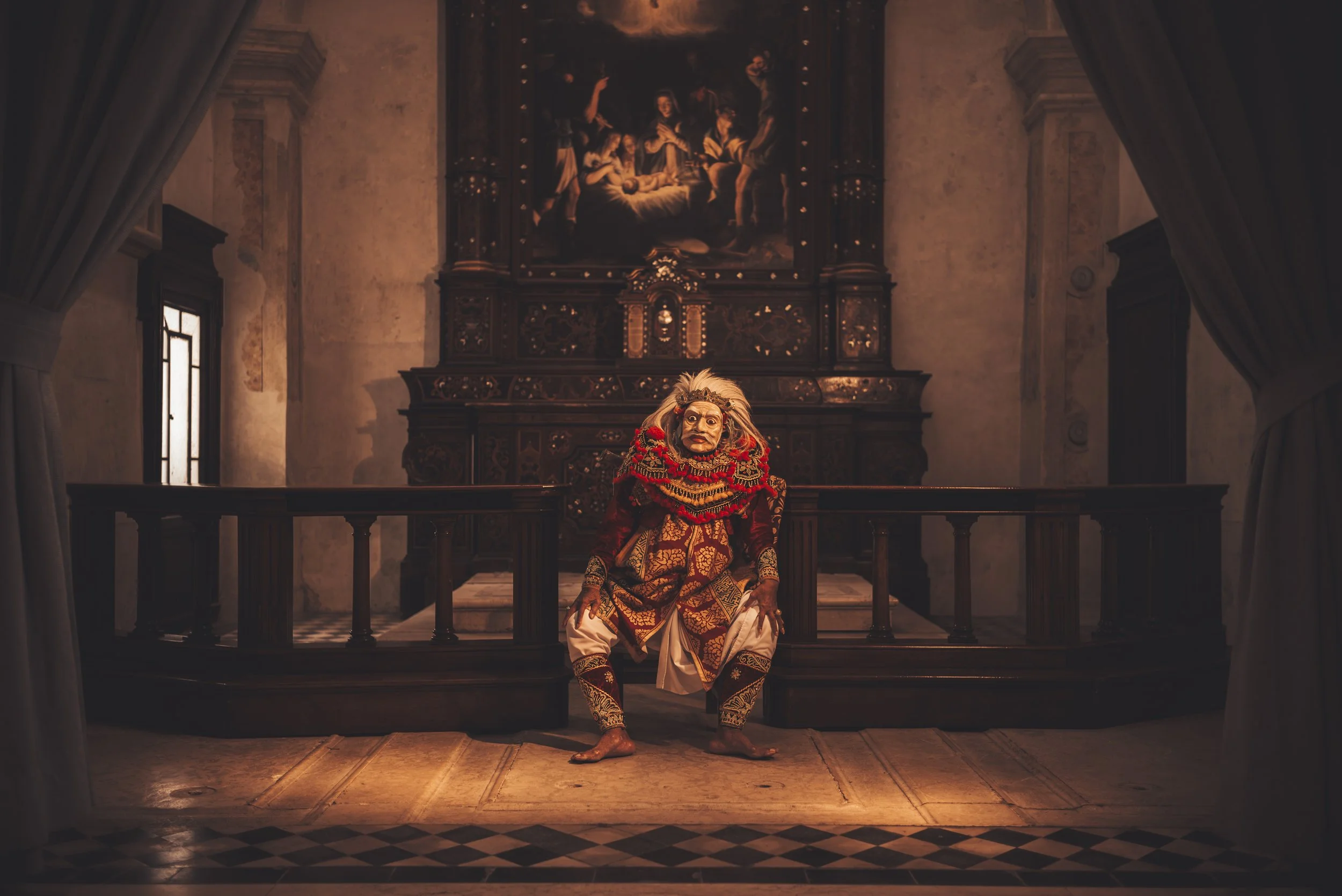 A performer in traditional mask and colorful costume crouches on stage in a room with ornate dark wood furniture and a religious painting behind. Curtains frame the scene, and sunlight streams through tall windows.
