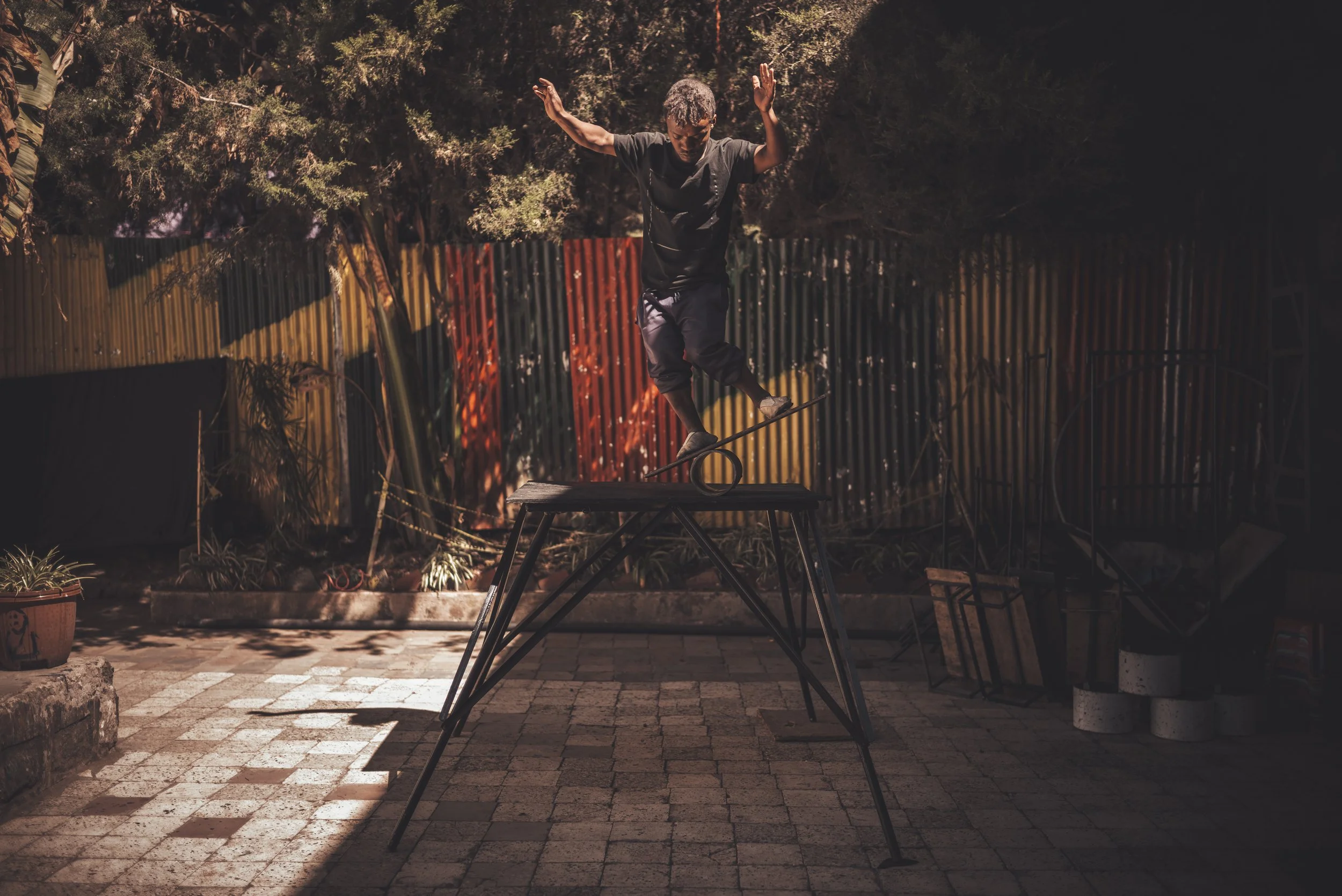 A young man balancing on a slackline set up between two elevated platforms in an outdoor area at dusk, surrounded by trees and a colorful metal fence.