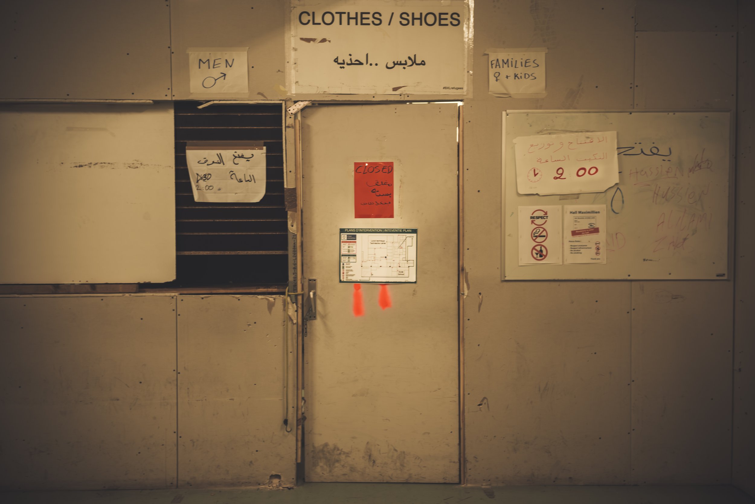 A wall with signs and notices indicating areas for clothes and shoes. There are labels for men, families, and a sign indicating the area is closed. The wall appears aged and slightly dirty, with handwritten and printed notices attached.