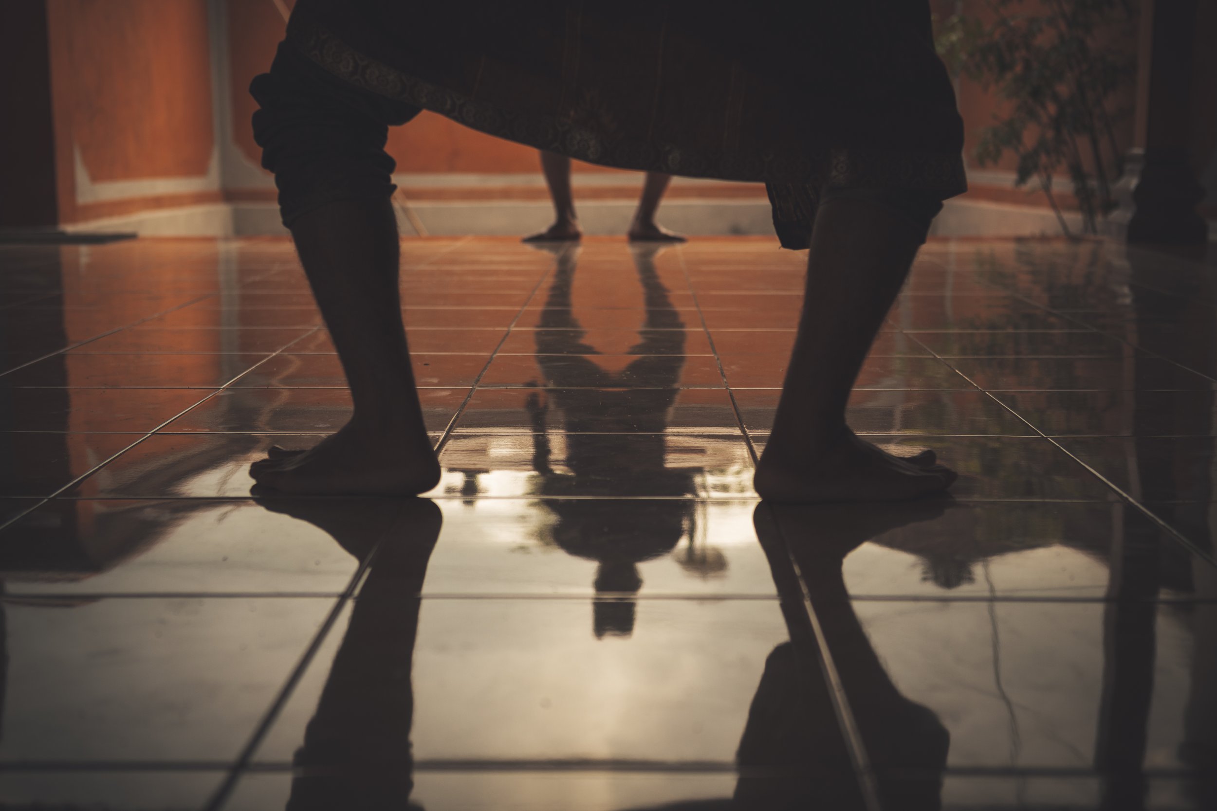 A person doing a handstand on a shiny tile floor, with their legs extended and feet flat on the ground, in a dimly lit room with warm tones and potted plant in the background.