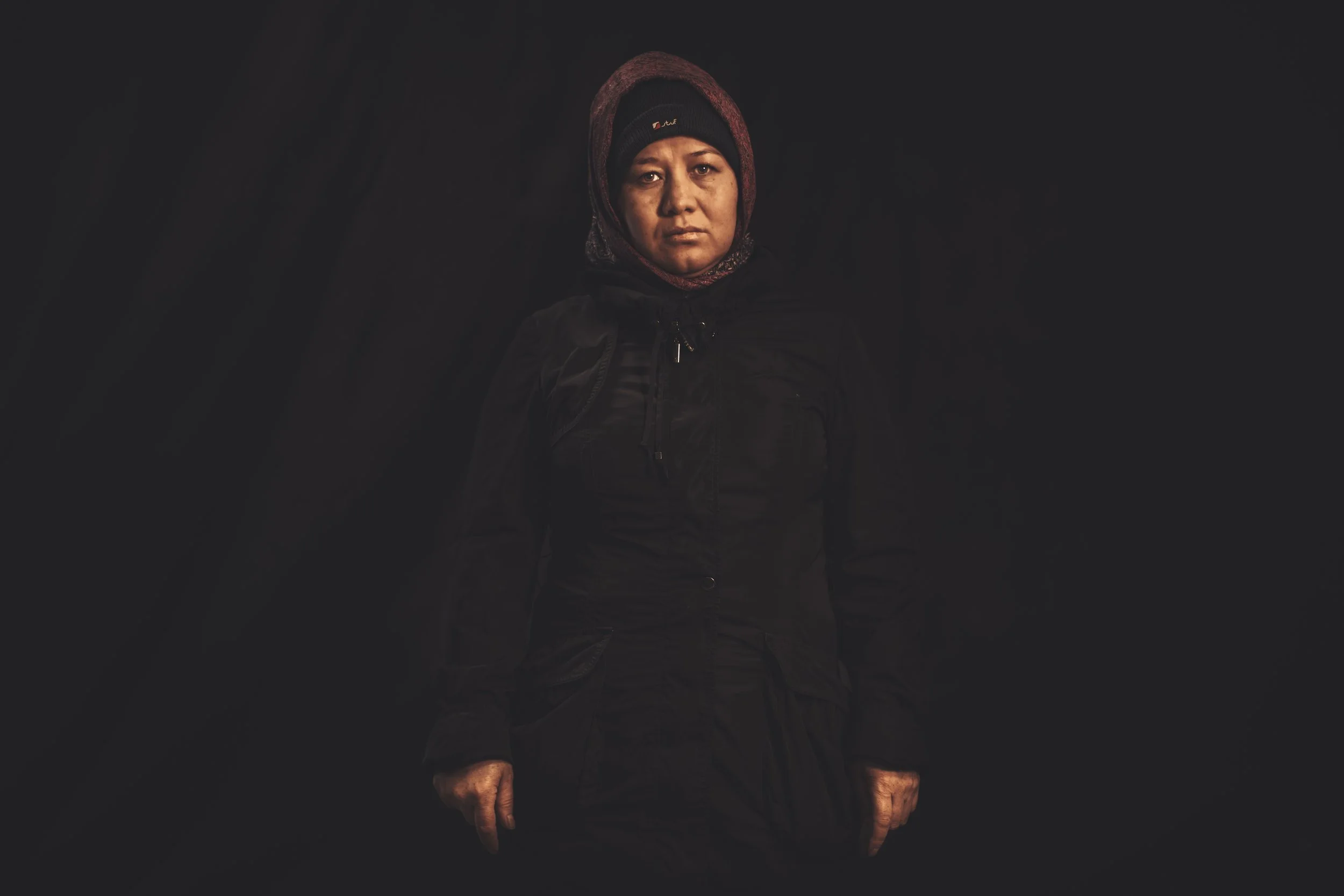 Portrait of a woman wearing a dark jacket and a beanie, standing against a dark background with serious expression.