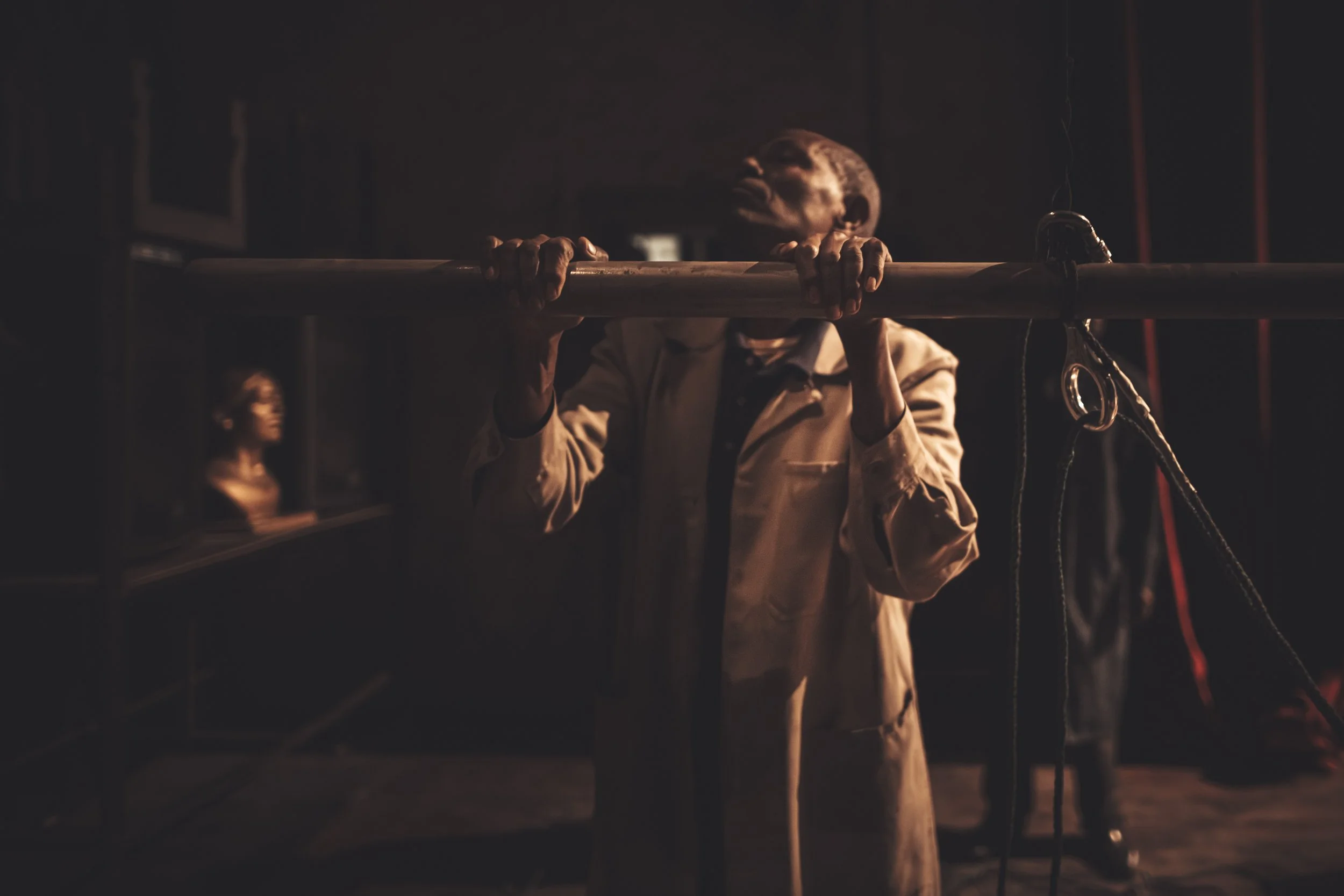 A man dressed in a beige suit holds onto a horizontal pole with both hands, with a serious expression on his face. In the background, there is a woman with glasses standing in front of a shelf with books in a dimly lit room.