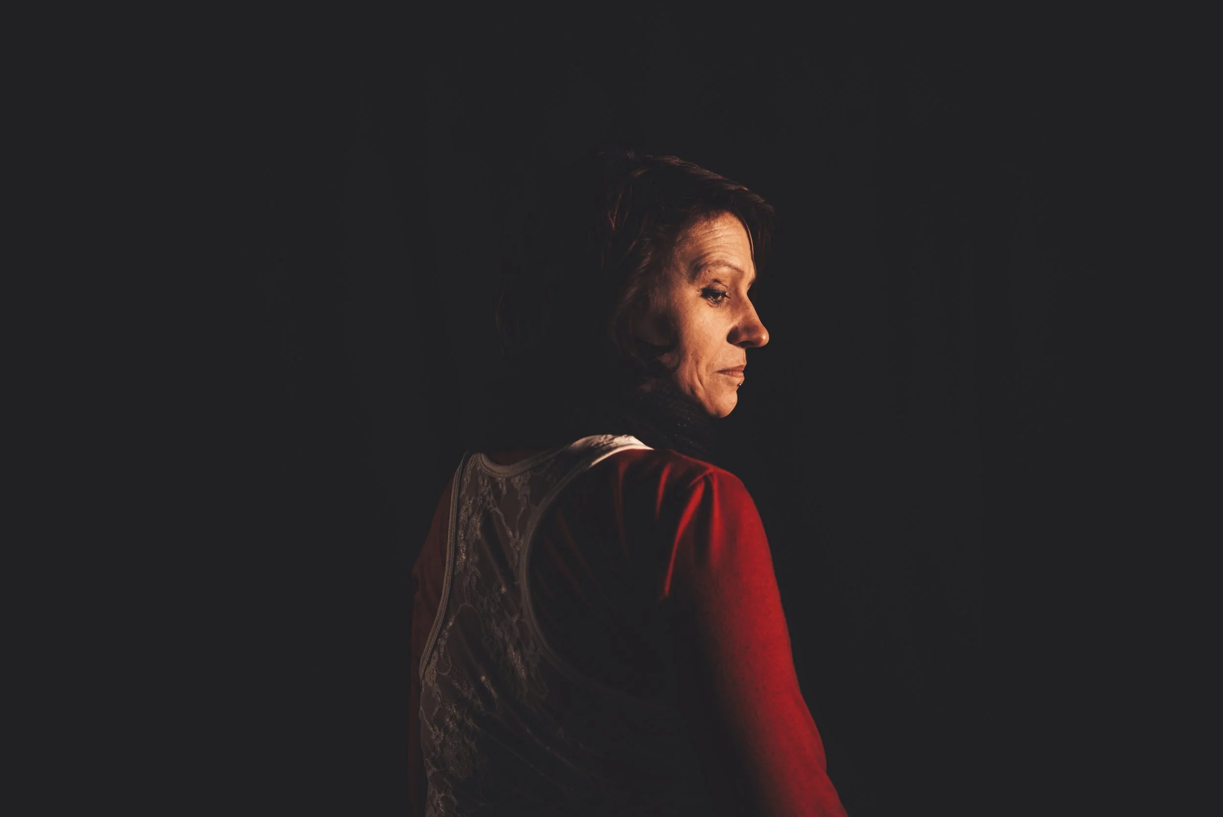 A woman with short hair, wearing a red and patterned top, looking downward in a dark setting with side lighting.