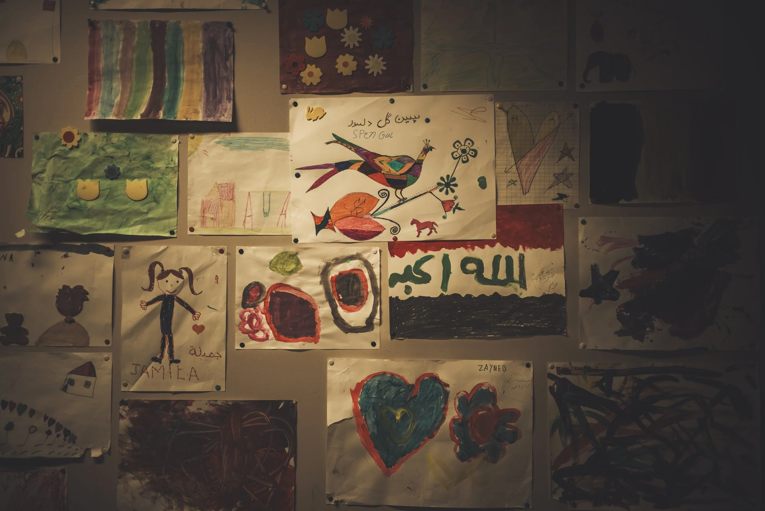 A collection of children's drawings and paintings on a wall, including colorful abstract art, flowers, a bird, hearts, a girl, and various shapes and symbols.
