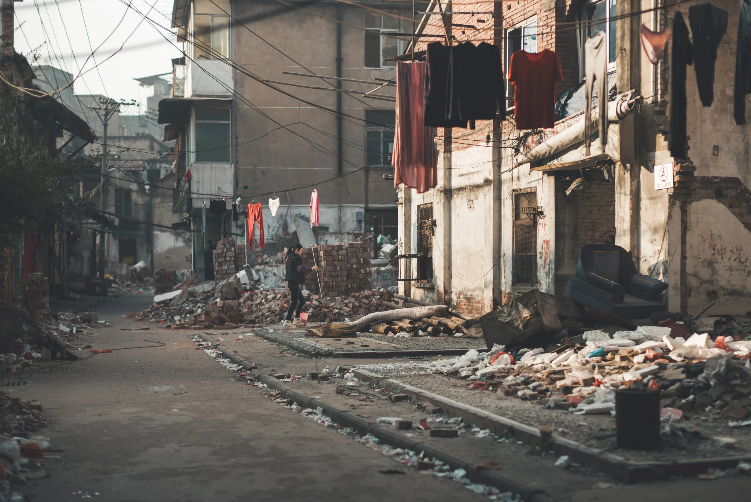 A woman sweeping debris in a damaged urban street with rubble, bricks, and broken infrastructure, likely after a disaster.