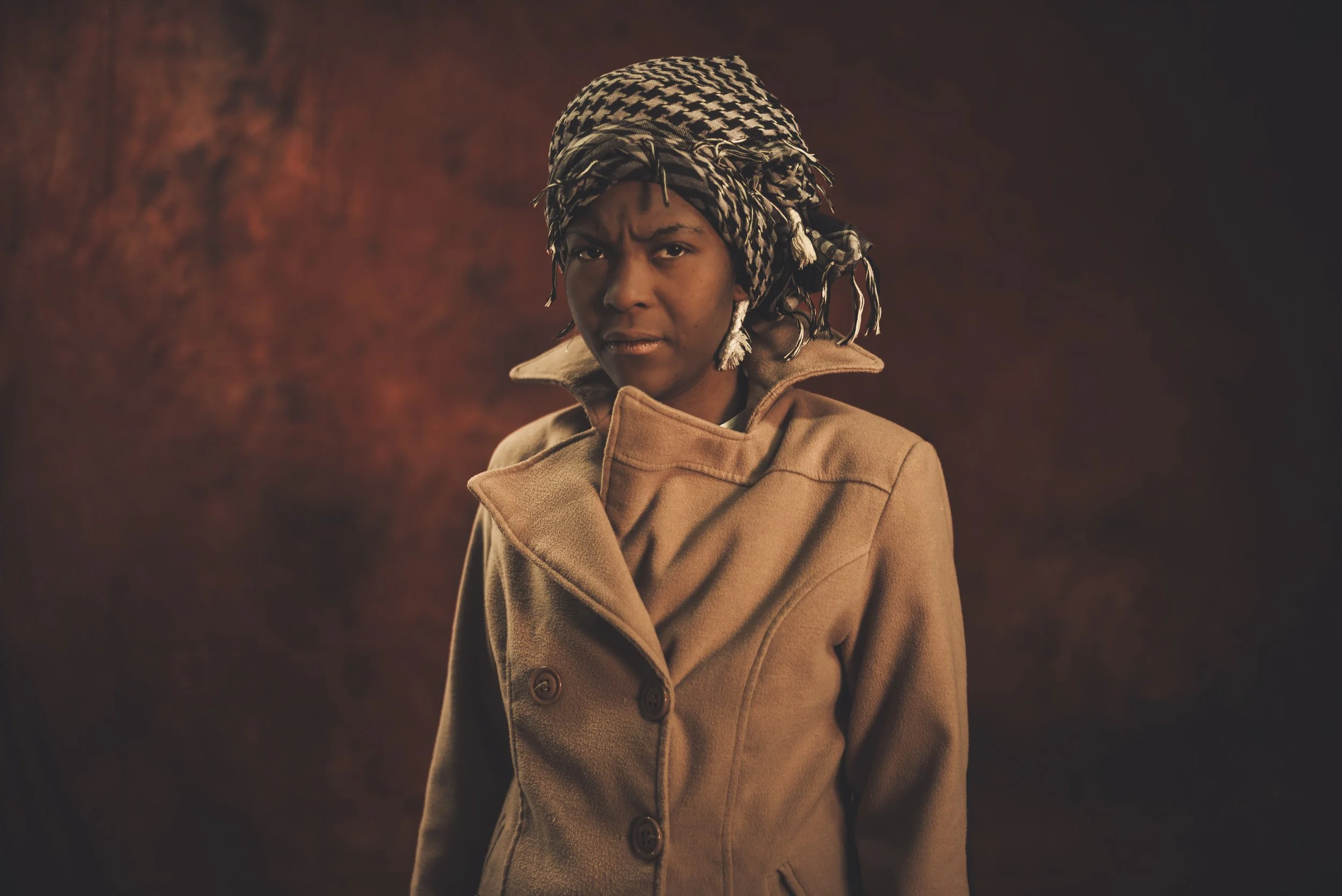 A young person with a tan coat and a patterned headscarf standing against a warm-colored background.