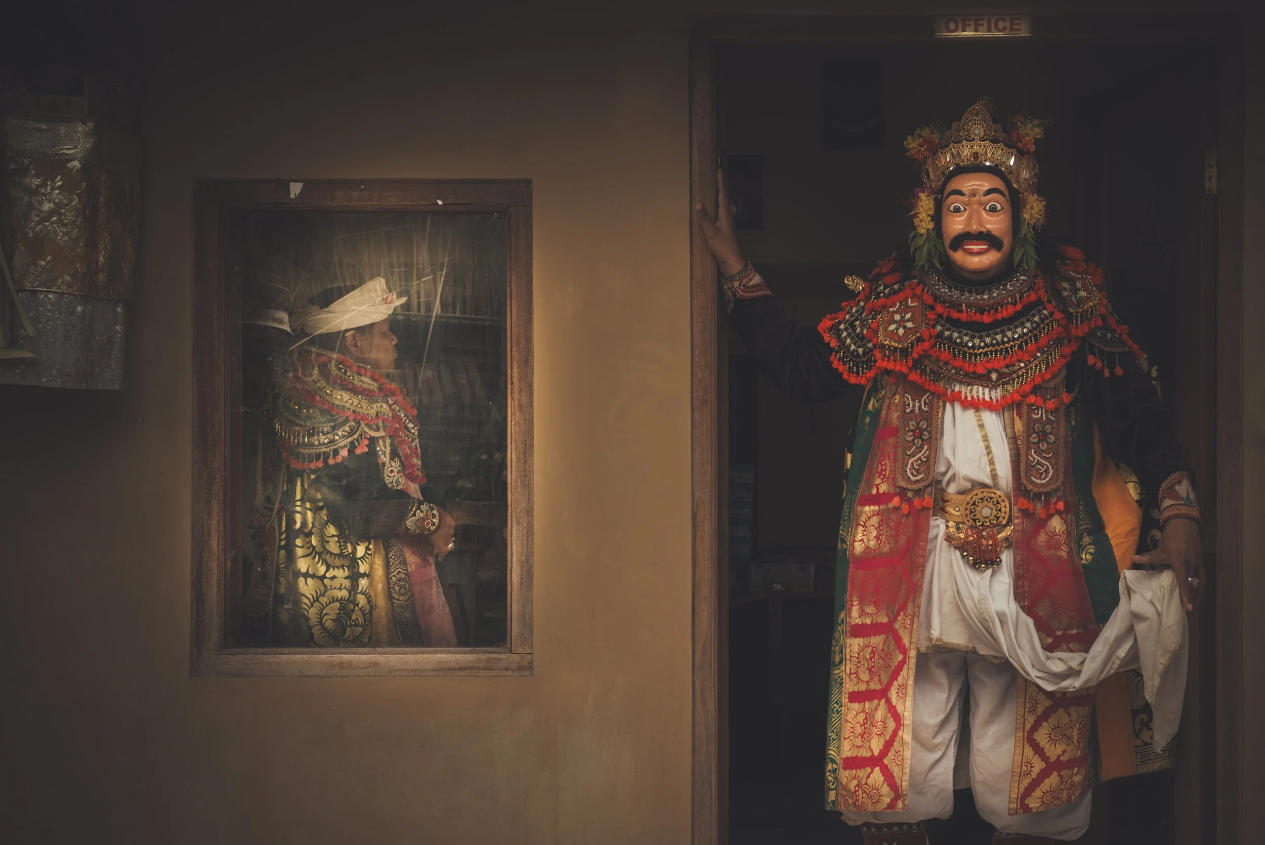 A person dressed in traditional elaborate colorful costume and a large mask, standing in a doorframe marked 'OFFICE'. Inside a window, another person in similar attire is visible, looking out.
