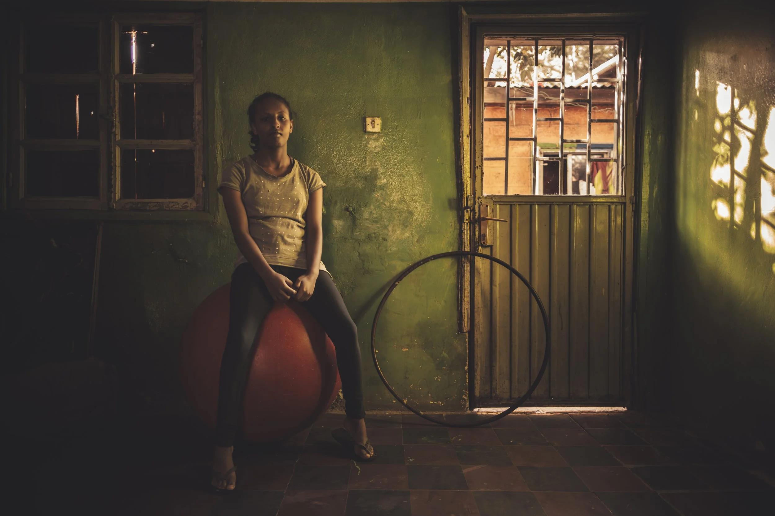 A young girl sitting on a red chair in a dimly lit room with green walls, with a door and window behind her. Light from outside illuminates part of her face and the room.