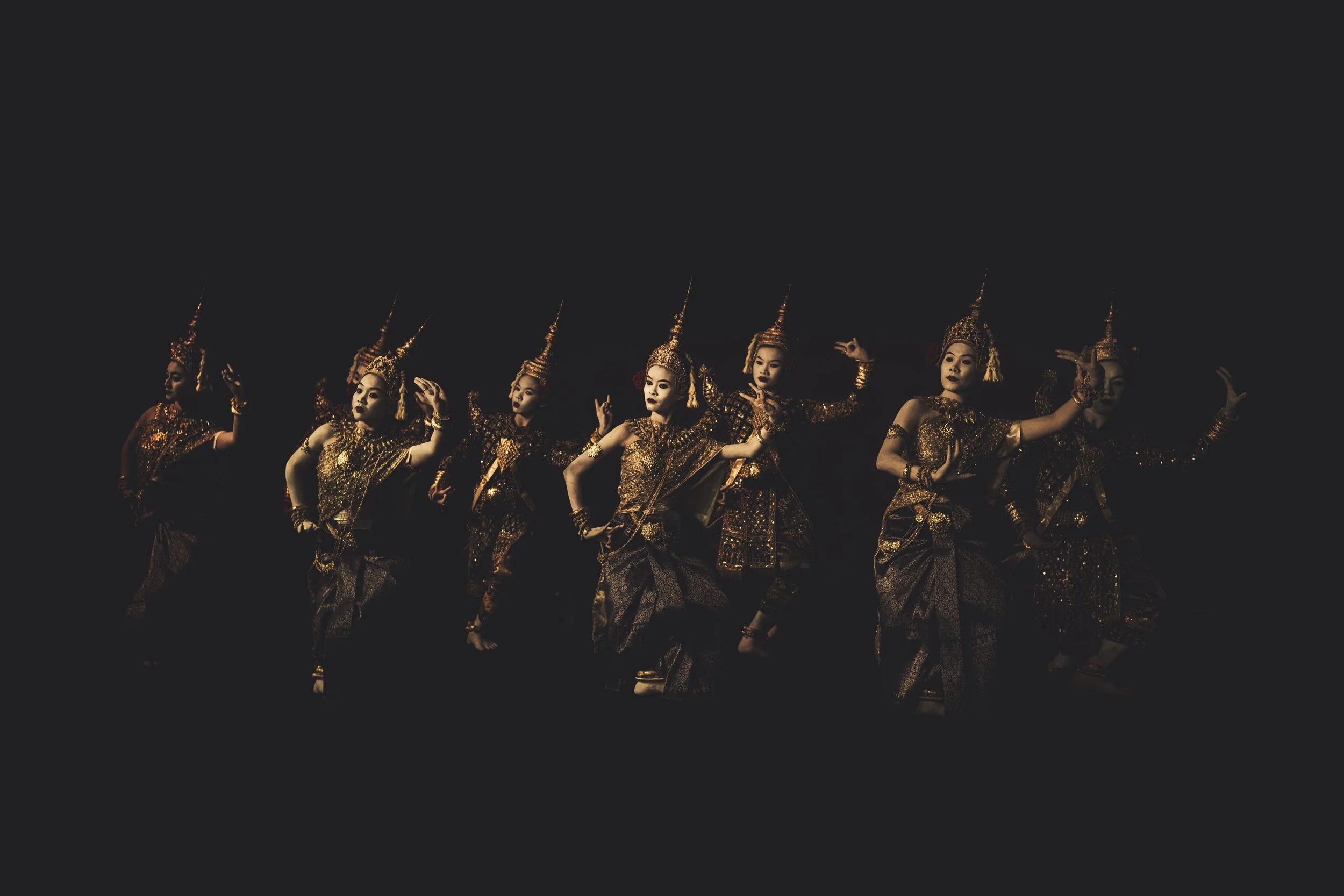 Seven women dressed in traditional gold and black costumes perform a dance on stage at night.