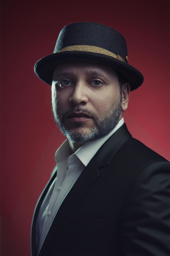 Man in a black suit, white shirt, and fedora hat against red background.