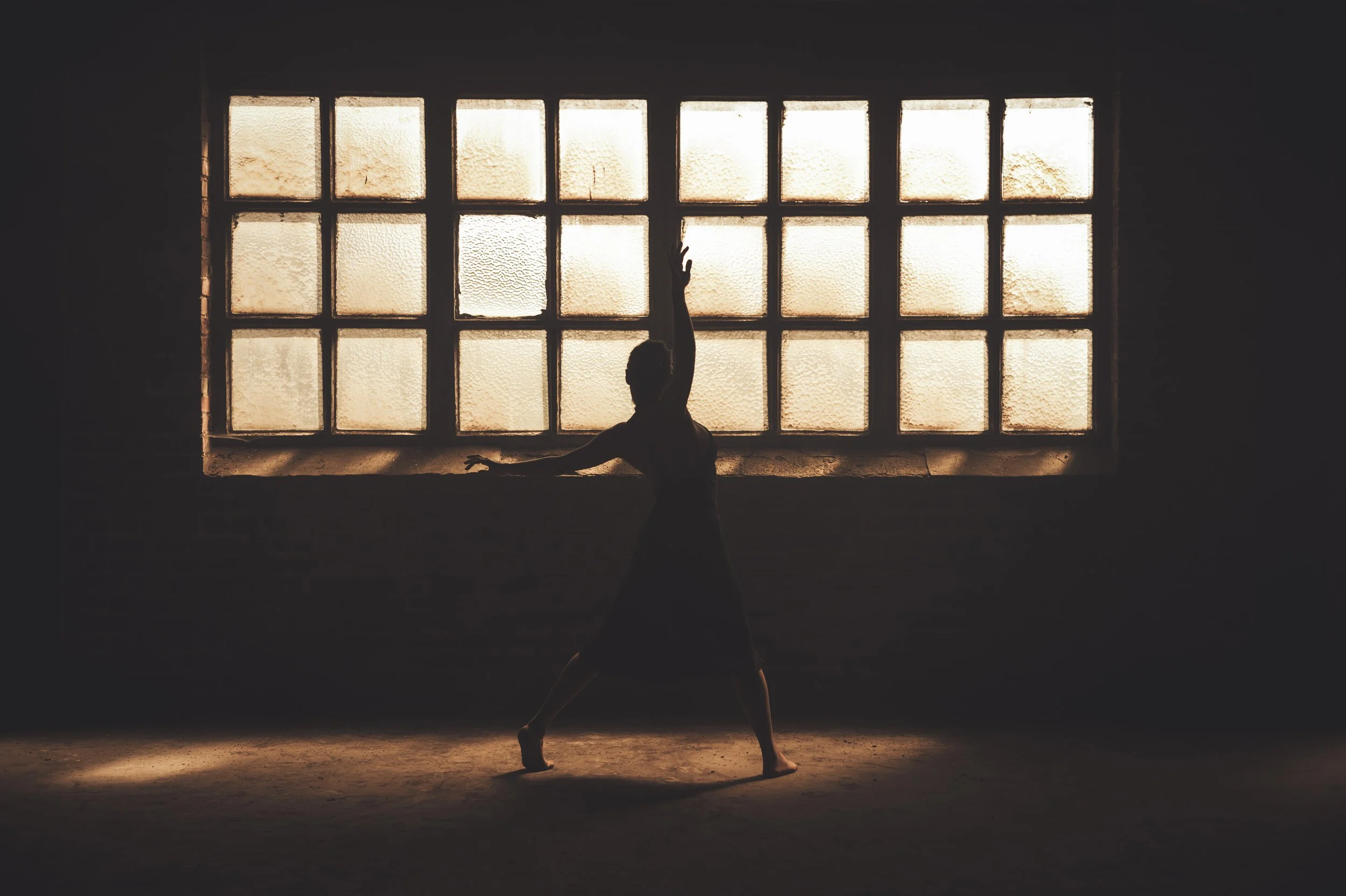 A silhouette of a ballet dancer practicing indoors in front of a large, textured glass window with light shining through.