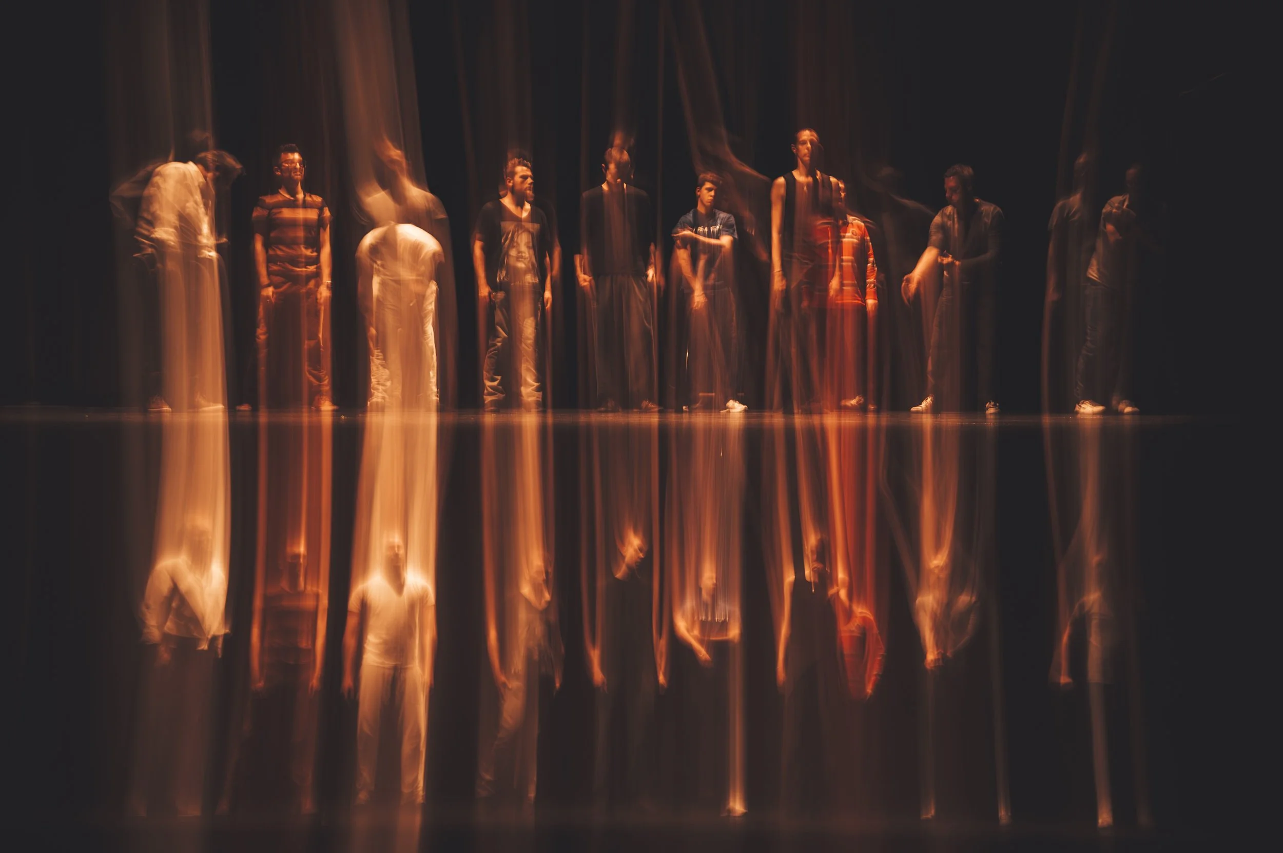 Fifteen people standing on a dark stage with streamers of orange and yellow light flowing downward from above.