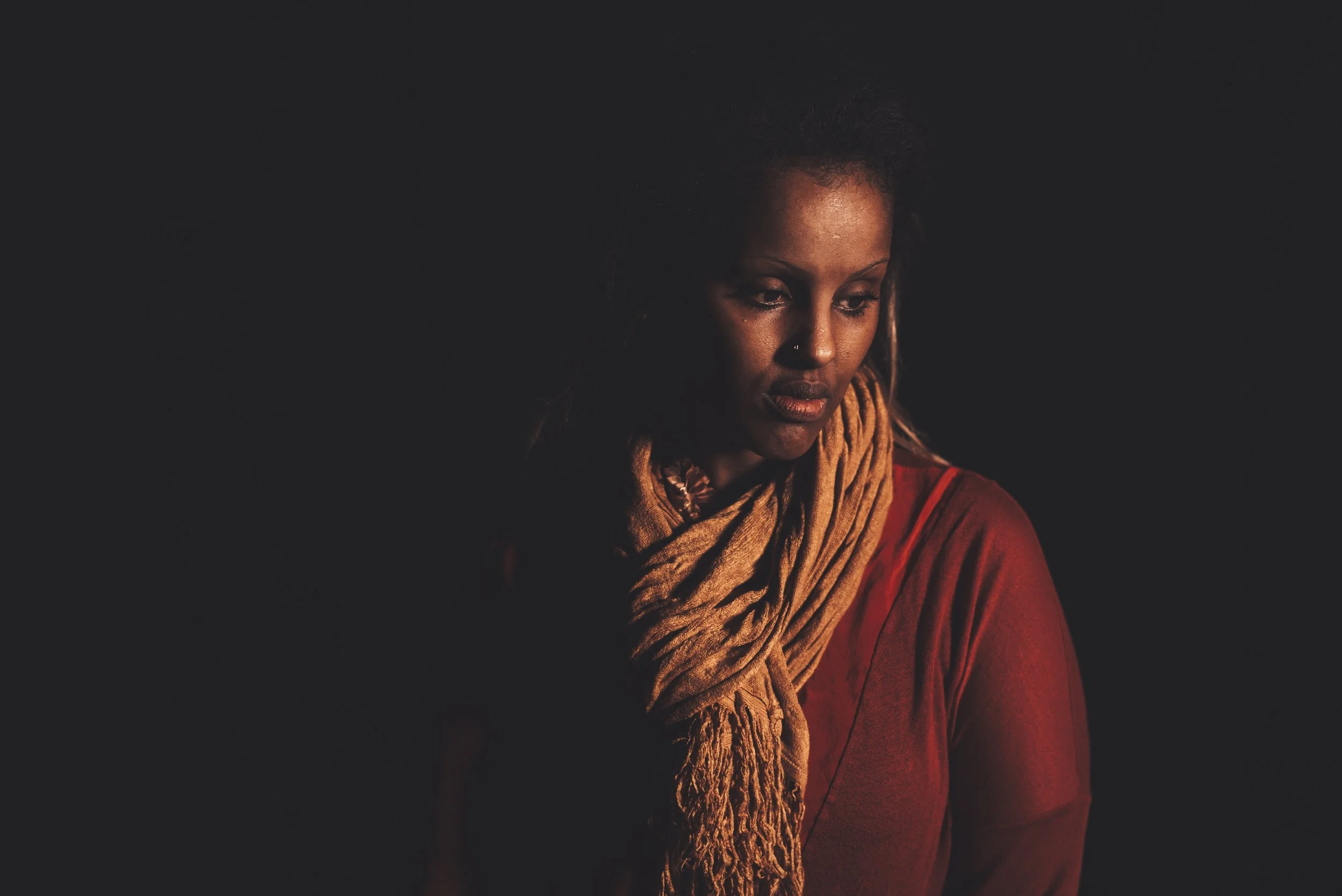A woman with dark skin and dreadlocks, wearing a red shirt and a beige scarf, looking down with a serious expression against a dark background.