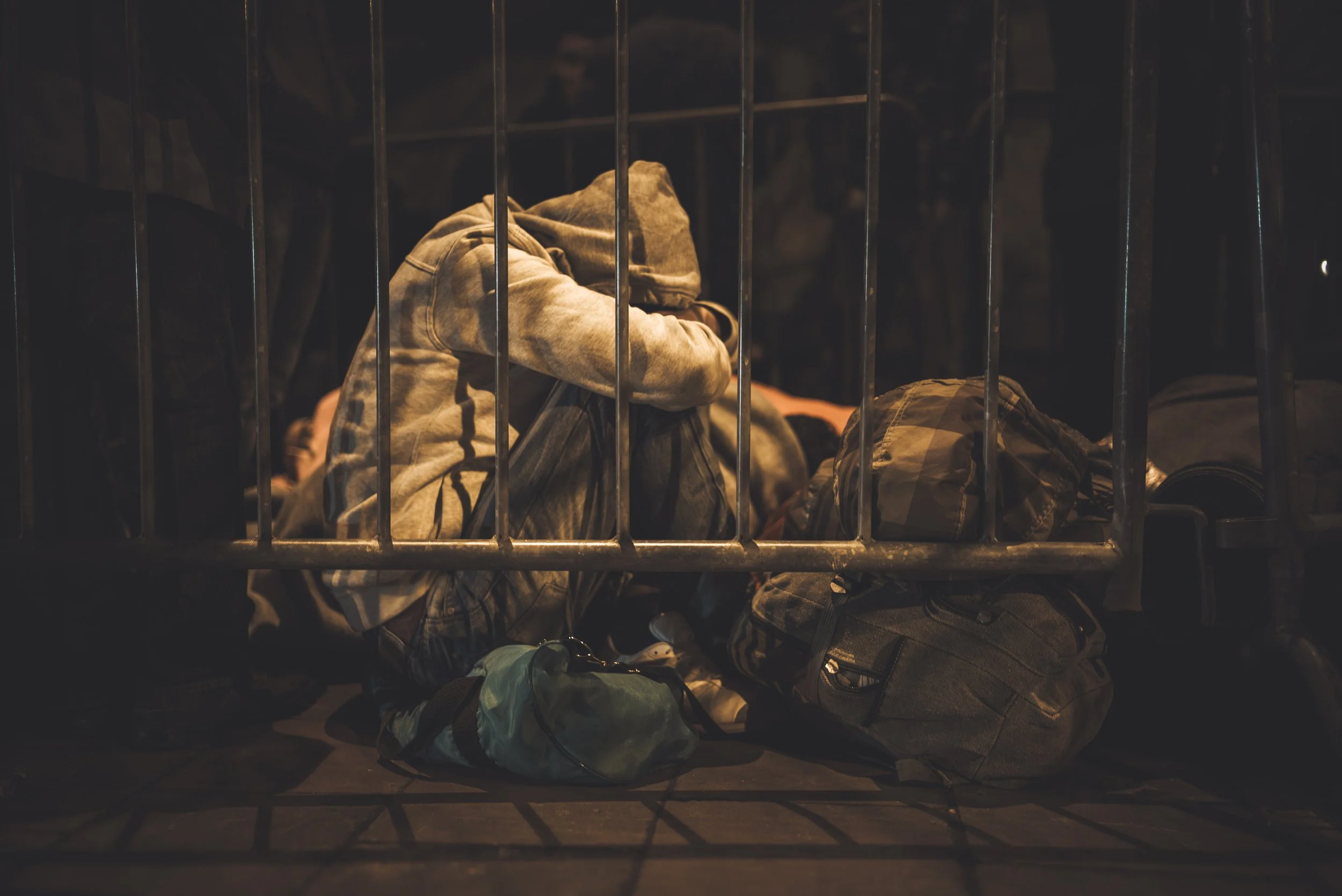 Person sitting on the floor behind bars, surrounded by backpacks and personal belongings, in a dimly lit area.
