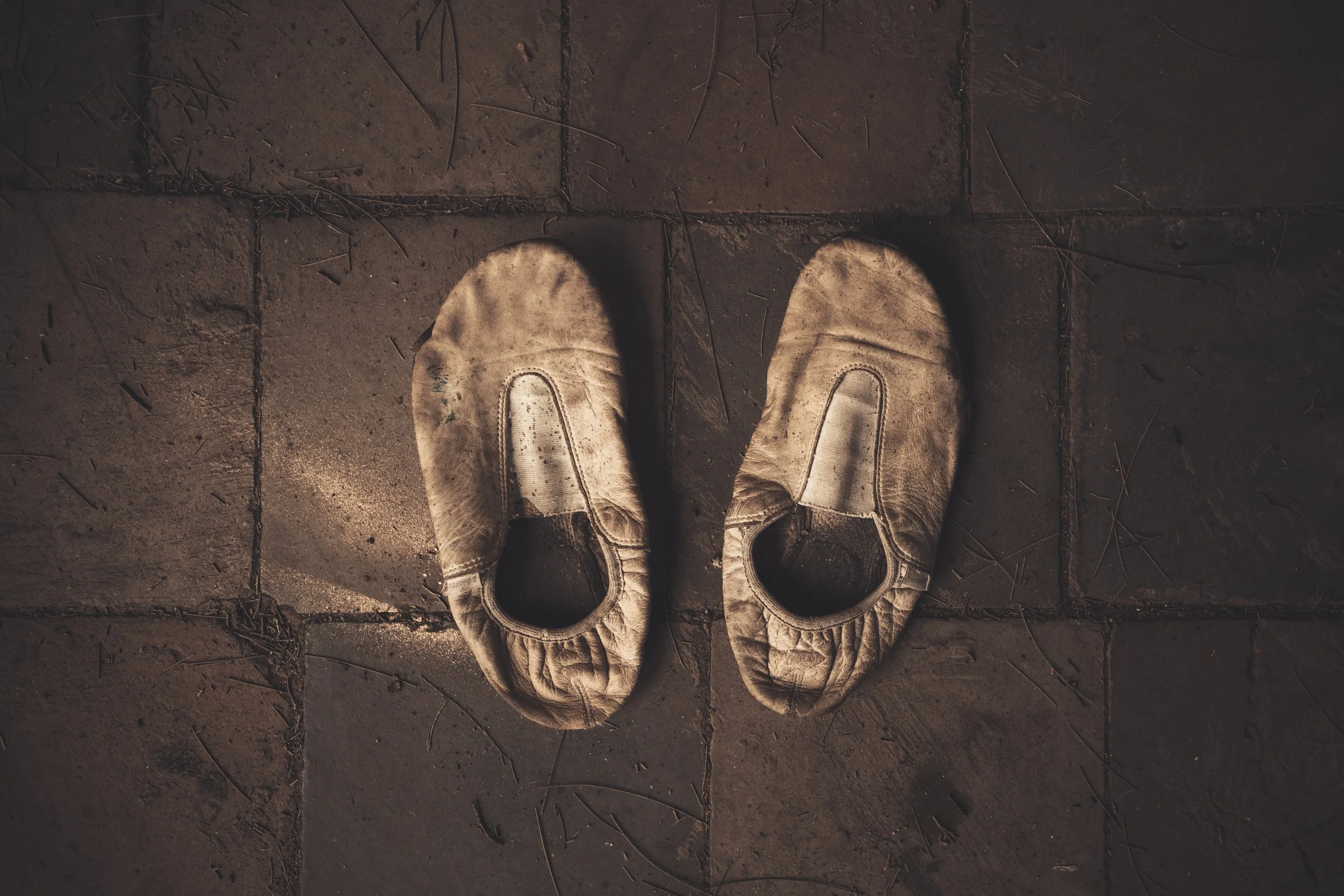 A pair of worn, white fabric sneakers with velcro straps, placed on a dimly lit stone or concrete floor with scattered small twigs and debris.