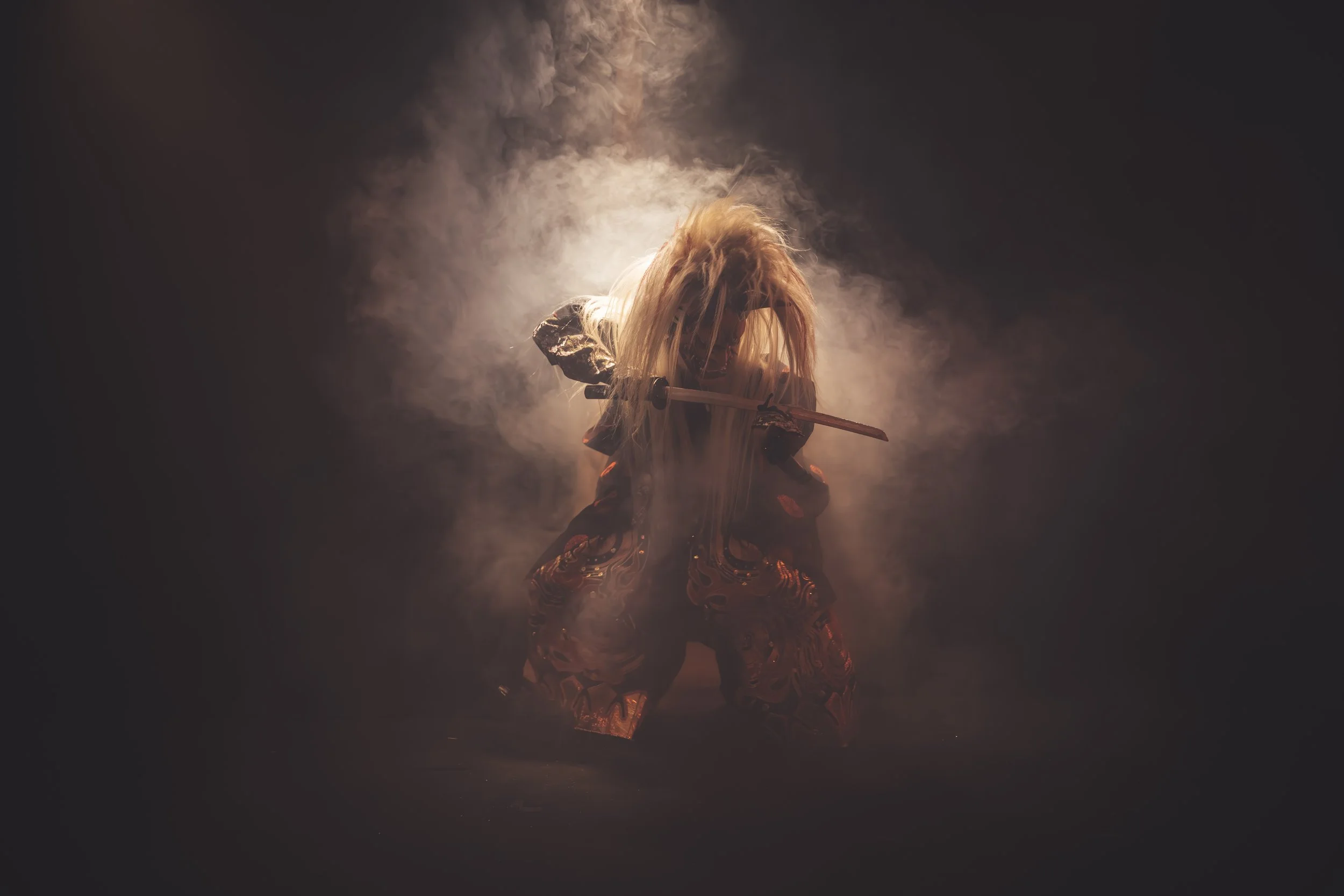 Person with long blonde hair wearing patterned clothing, holding a sword, surrounded by smoke or mist, with a dark background and a bright light source behind them.