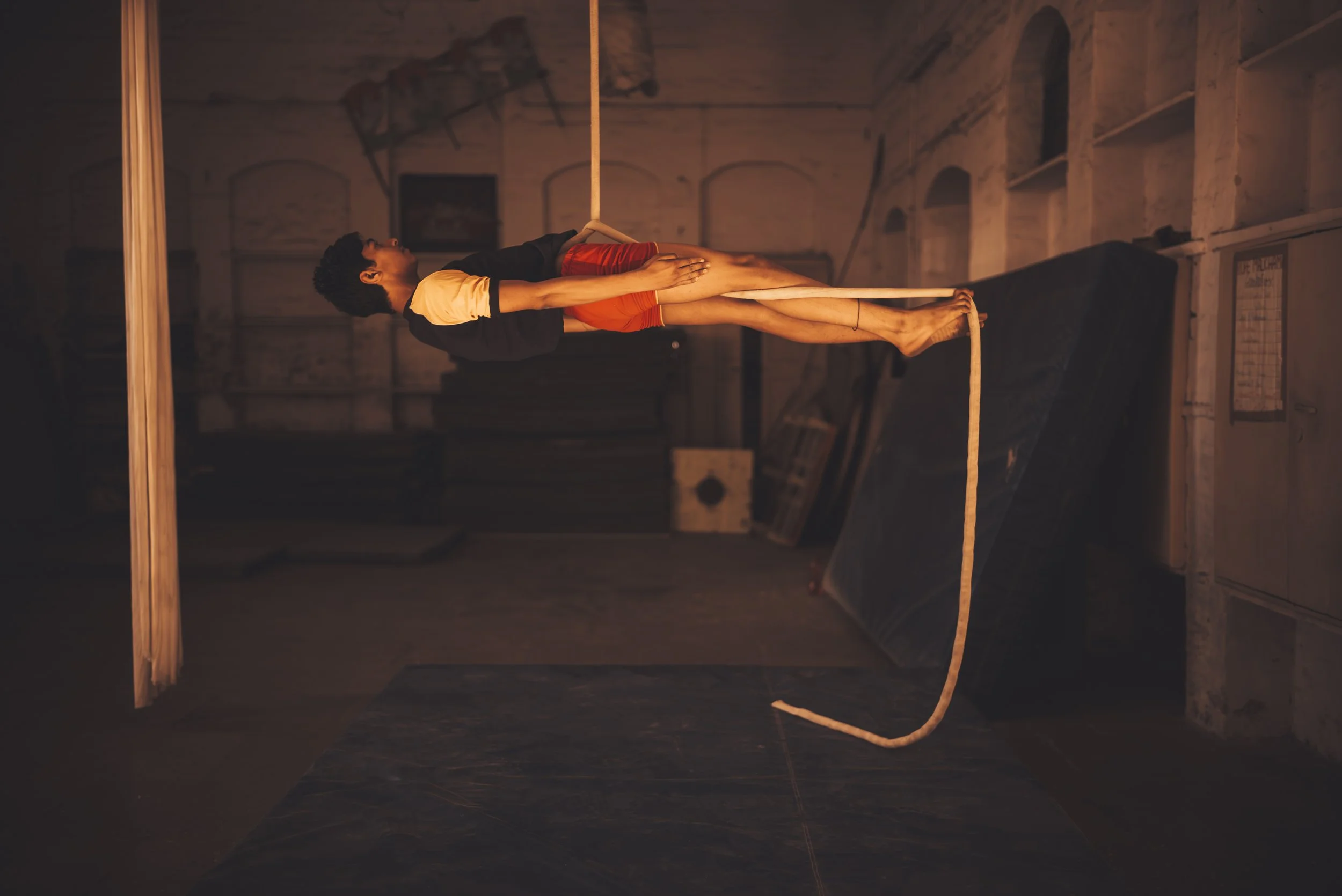 A person performing an acrobatic pose on a hanging trapeze in a dimly lit gym or studio with brick walls and wooden shelving.