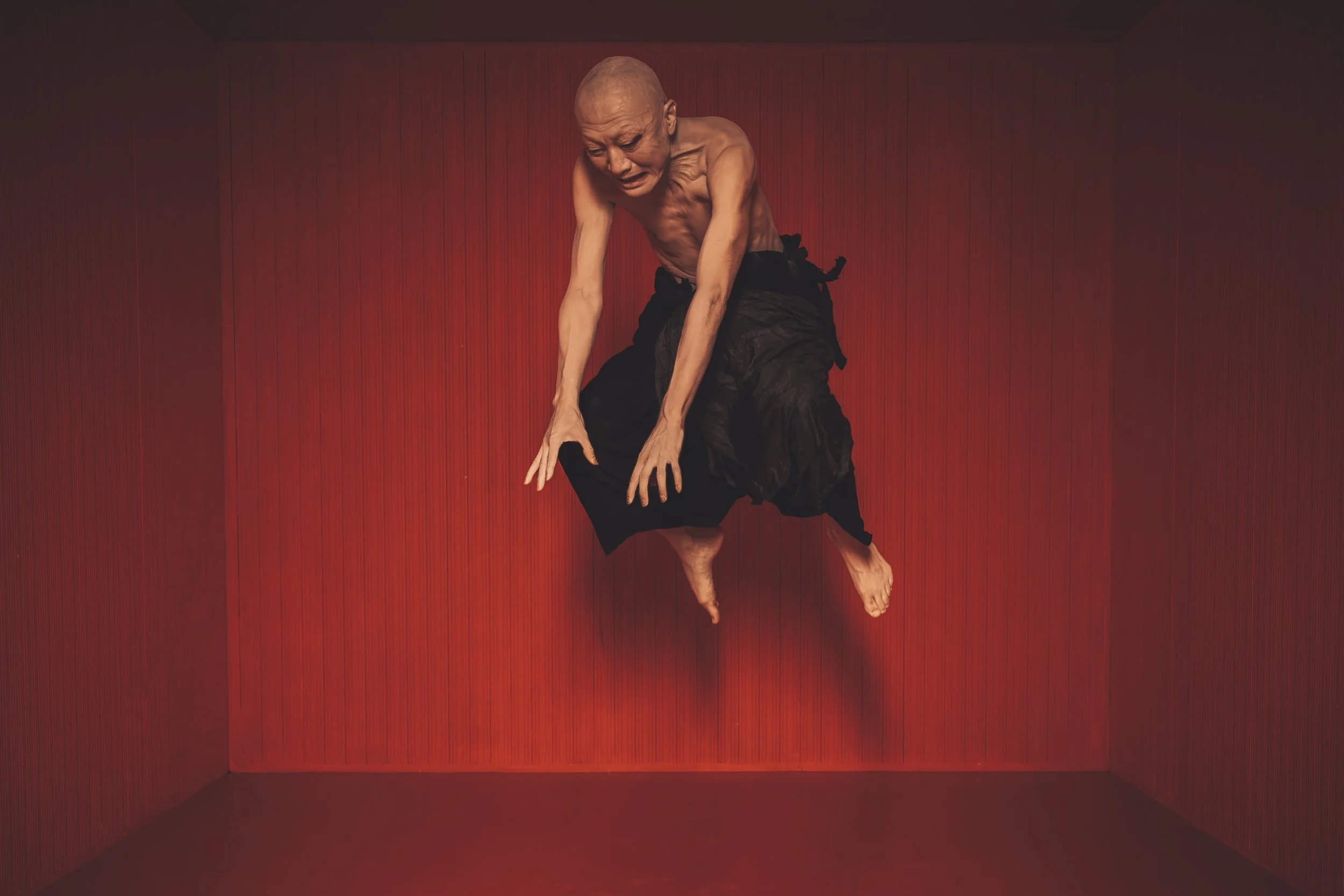 An elderly man with a shaved head and a muscular build is seen performing a yoga pose against a red wall. He is wearing black pants and is balancing on one foot with his other leg crossed over his standing leg. His hands are resting on his bent knee 