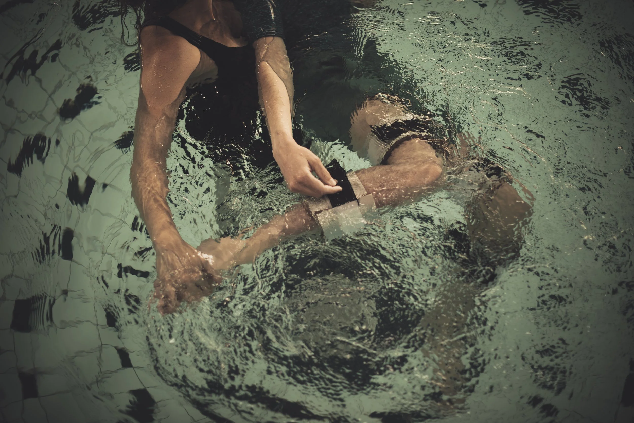 Two women in a swimming pool, one assisting the other with a wrist strap or bracelet.