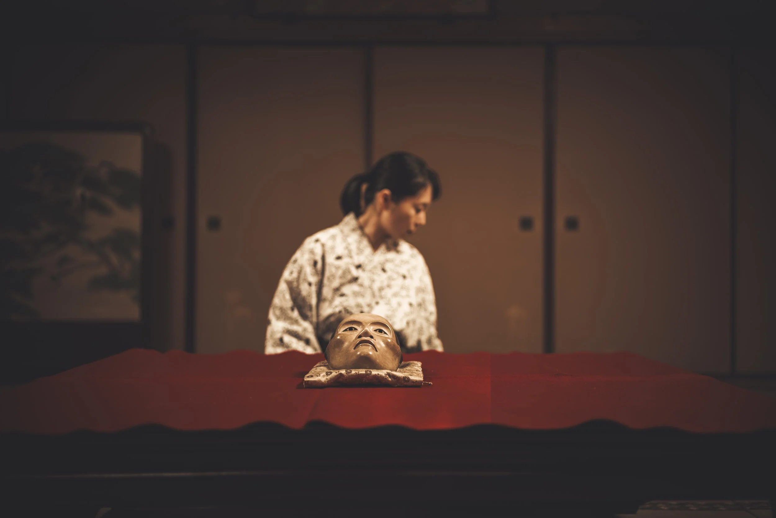 A woman dressed in traditional Japanese clothing stands behind a table covered with a red cloth, looking down at a mask resembling a human face placed in front of her. The background features a wooden wall with sliding doors and a painting of a lands