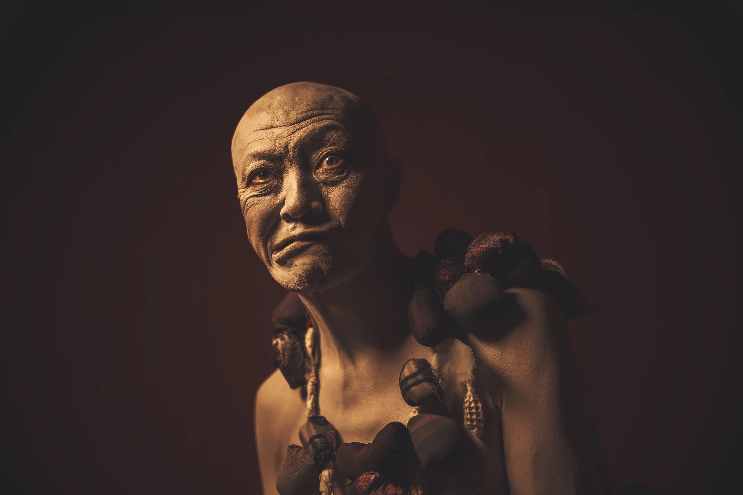 A person with a bald head and intense expression, wearing dark gloves and necklaces, illuminated by dramatic lighting against a dark background.