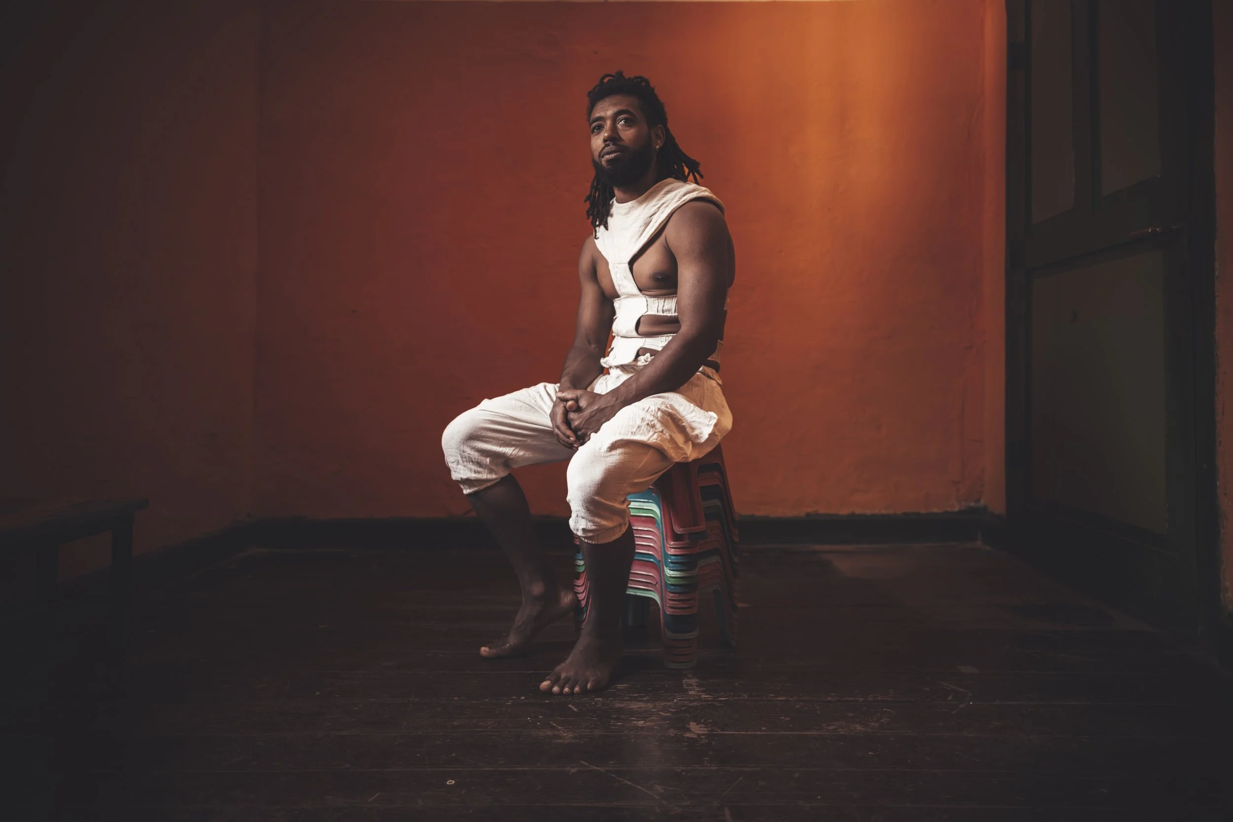 A man with a beard and dreadlocks sitting on a stack of plastic chairs against an orange-red wall, wearing beige pants and a torn white sleeveless shirt.