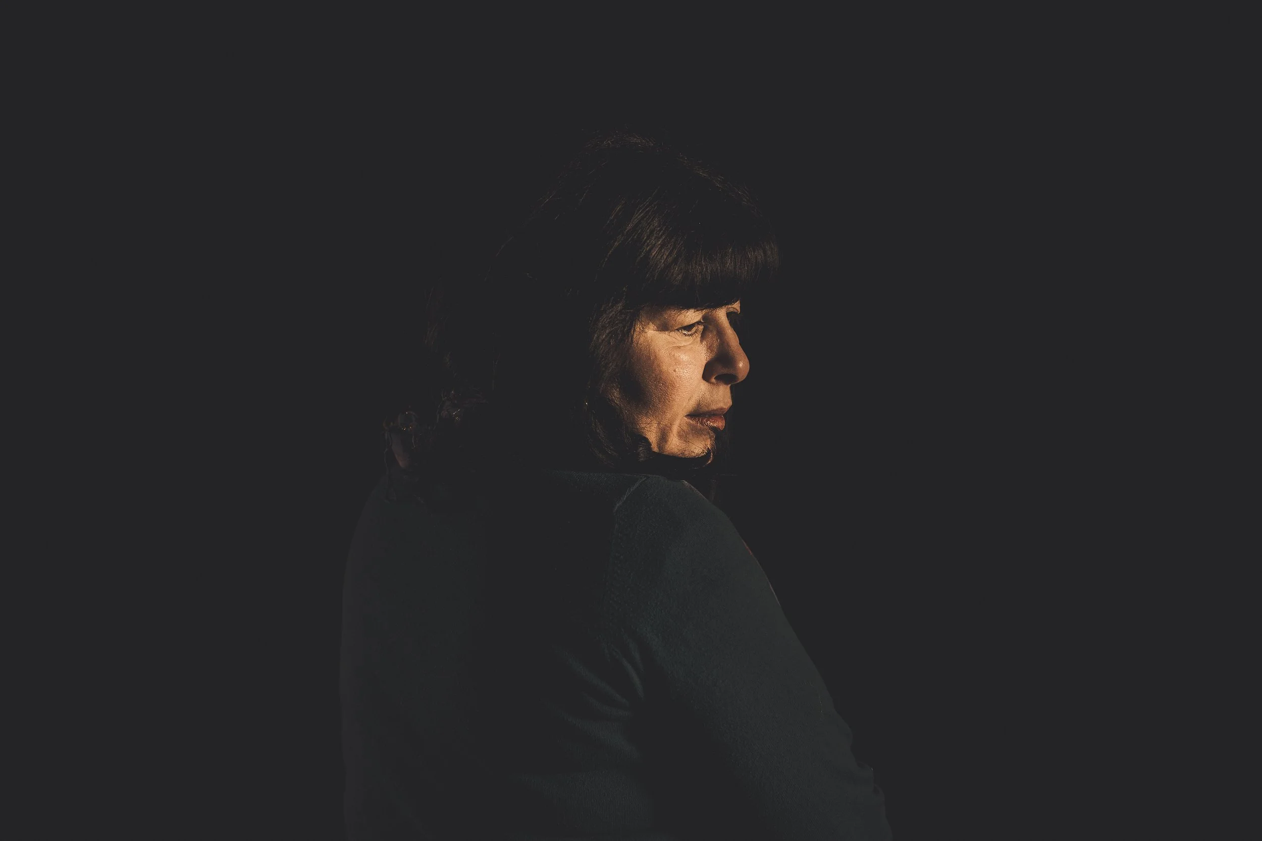 Profile of a woman with dark hair and bangs, facing sideways against a dark background.
