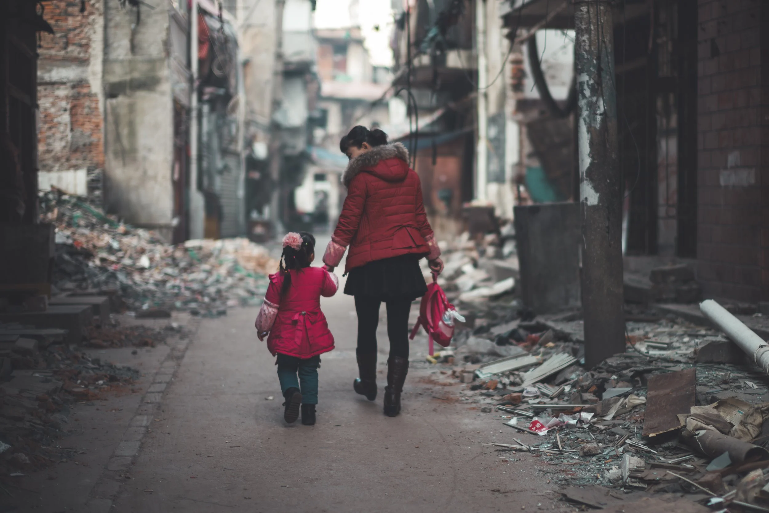 A woman and a girl in red jackets walking through a destroyed alley with debris and damaged buildings.