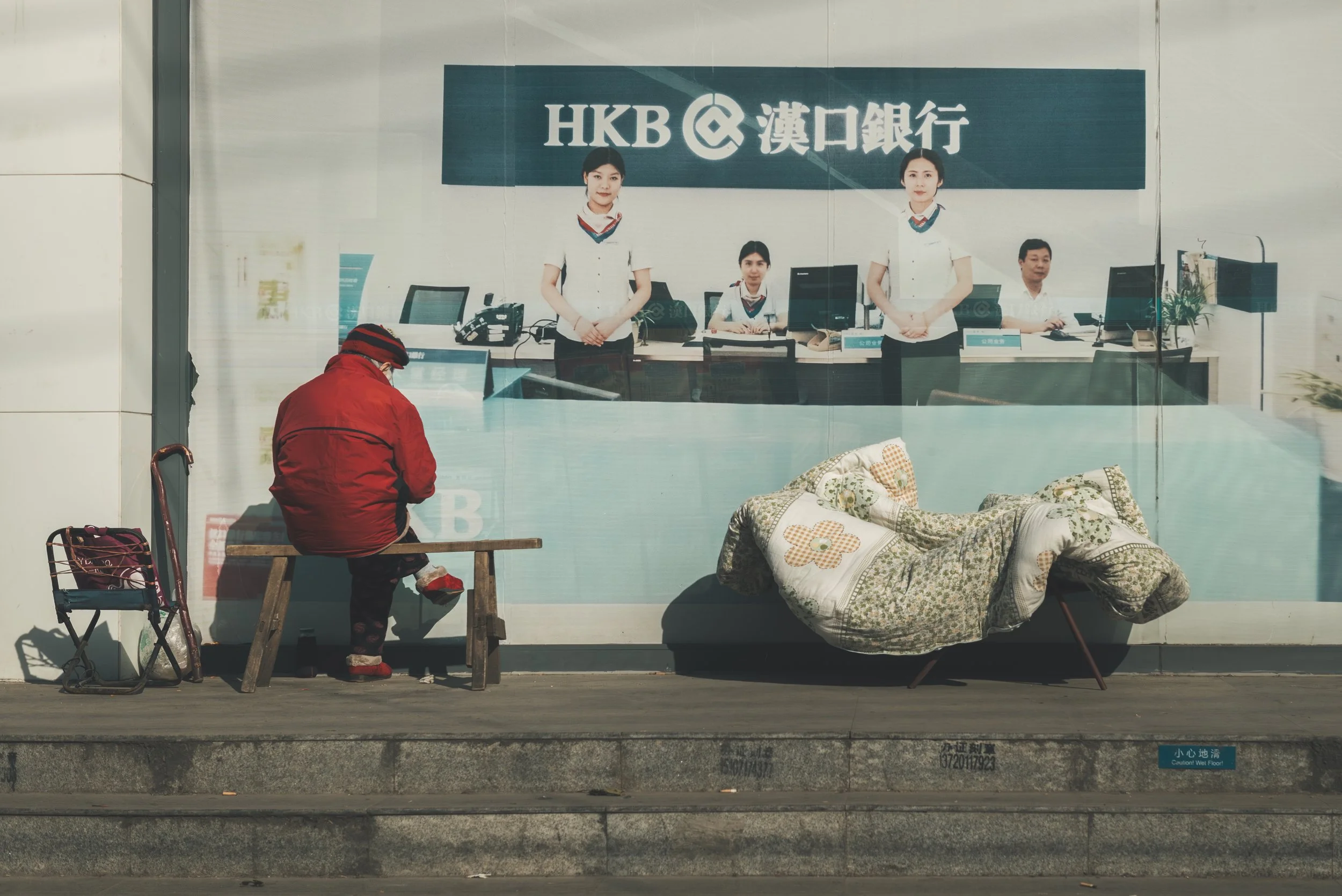 View of the outside of a bank with a glass window showing bank staff and a man waiting outside. The bank sign reads 'HKB' with Chinese characters. Inside, four staff members are sitting or standing at the counter, wearing uniforms. Outside, a man in 